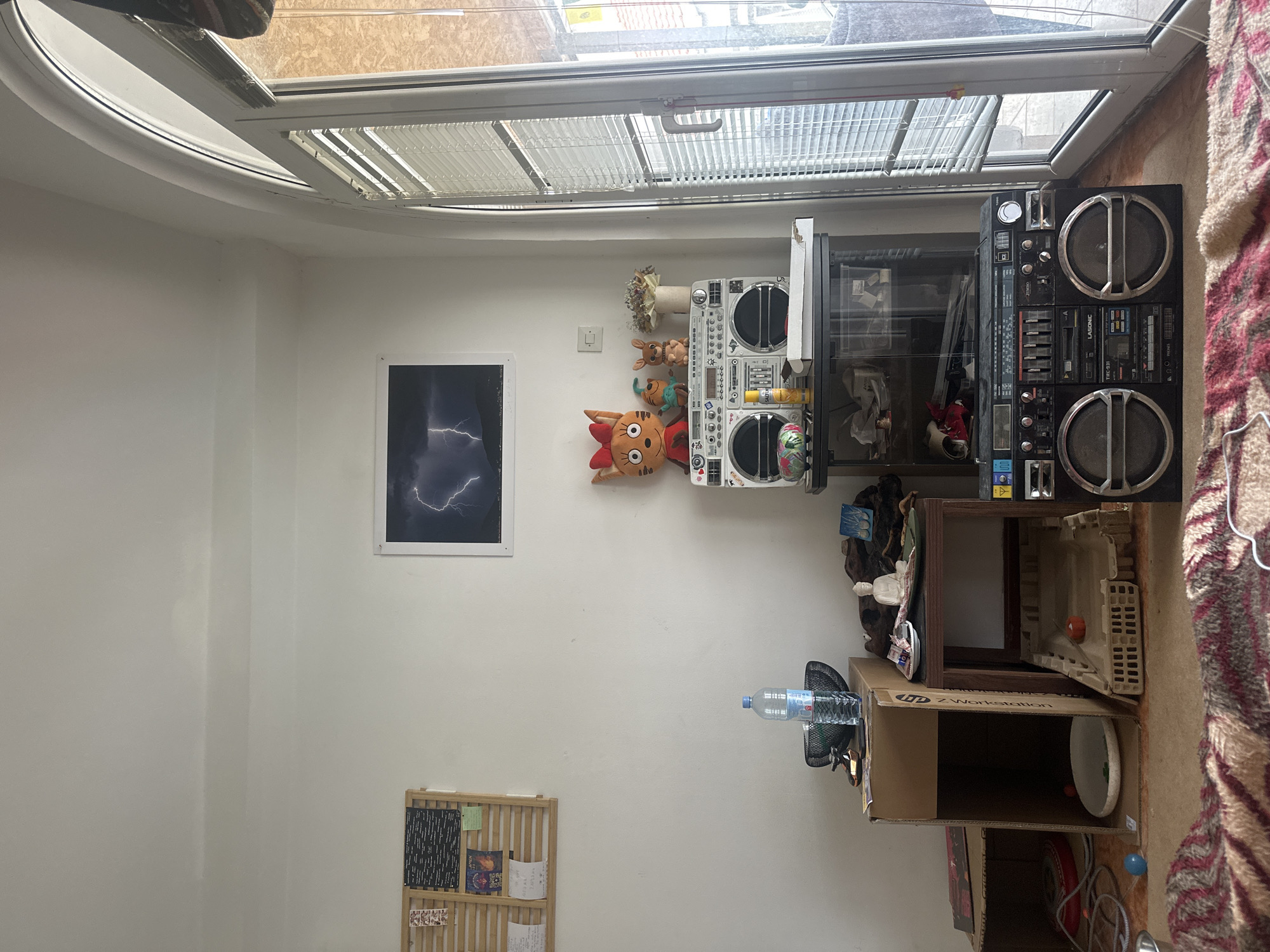 The image shows an interior space with white walls and a window running along the top of the frame. Under the window are a boombox and a smaller stereo system mounted on the wall. A framed picture hangs on the wall, depicting a lightning storm. Below the picture is a wooden shelf containing books and papers. Near the boombox is a cardboard box marked "HP Z Workstation". A water bottle is placed horizontally near the box. The image does not provide any information about the location of the room in Budva, Montenegro.