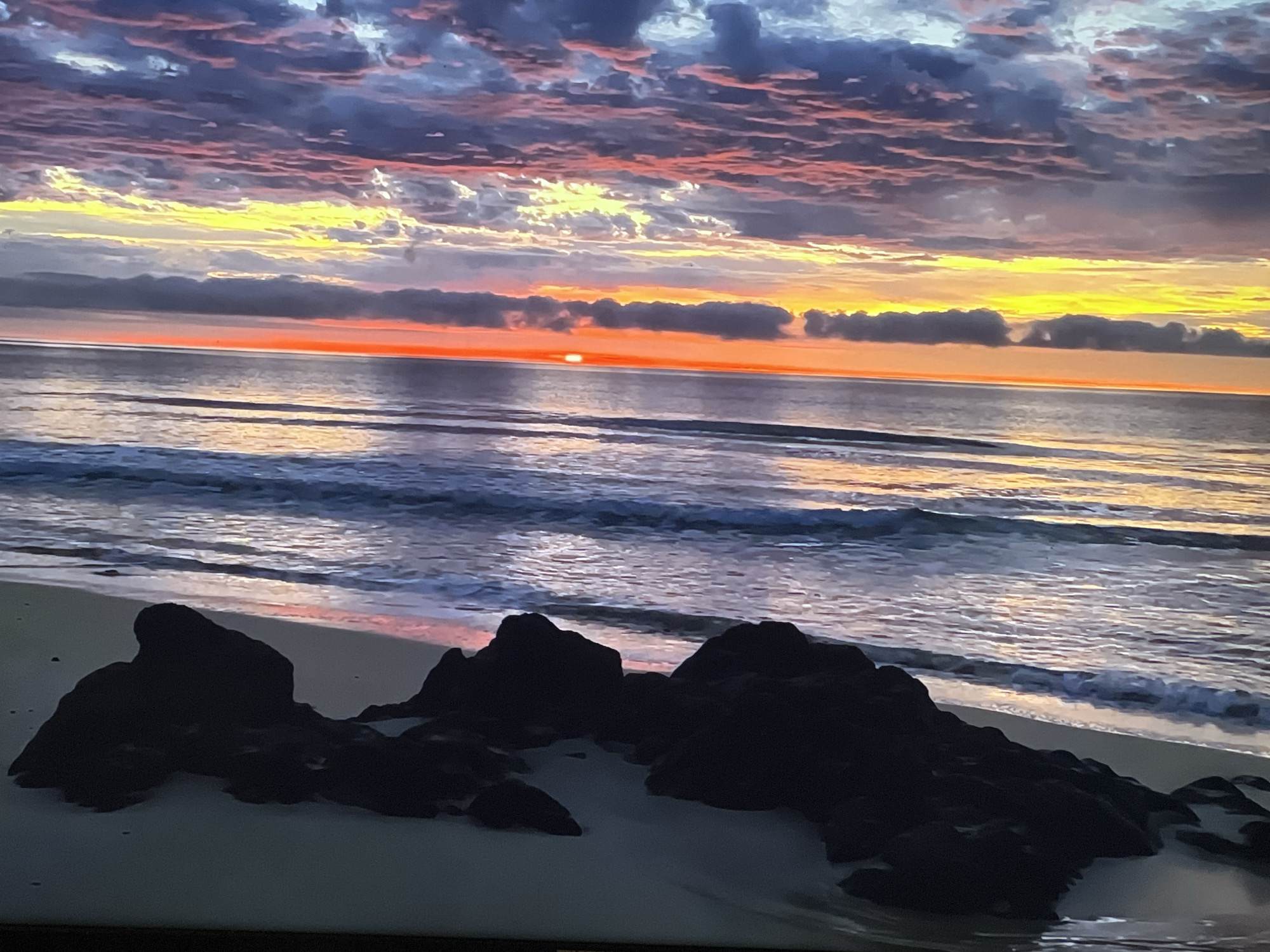 This media file shows a serene ocean sunset. The sky is ablaze with vibrant colors, transitioning from deep blues and purples at the top to fiery oranges and yellows near the horizon. Wispy, textured clouds are scattered across the sky, catching the light and adding depth to the scene.
The ocean is calm, with gentle waves rolling towards the shore. The water reflects the colors of the sunset, creating a shimmering effect. In the foreground, dark, silhouetted rocks are scattered on a sandy beach. The overall mood is peaceful and tranquil, evoking a sense of natural beauty and awe.
The time of day is clearly sunset, as indicated by the low position of the sun and the warm, vibrant colors in the sky. The weather appears to be clear and calm, contributing to the peaceful atmosphere. There are no people or visible text in the image. The setting is a beach with rocks, likely a coastal area. Given the location context of Sofia, Bulgaria, which is inland, this image likely depicts a coastal scene from elsewhere, possibly a vacation or a place with a different geographical setting.