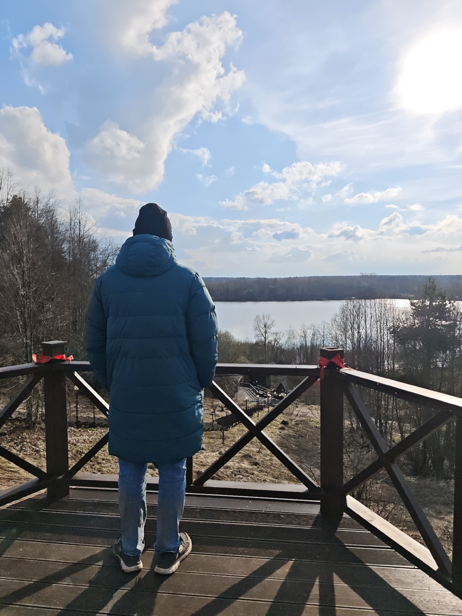 A person wearing a black beanie, a blue puffy jacket, and jeans stands on a wooden platform, looking out over a body of water. The platform is enclosed by a wooden railing with a decorative X pattern. Red ribbons are tied to the posts of the railing on either side of the person. Beyond the railing and platform is a view of trees and a lake, reflecting the sunlight. The sky above is blue with scattered white clouds and a bright sun. The scene appears to be Жестылево, Russia, as indicated by the geotag.