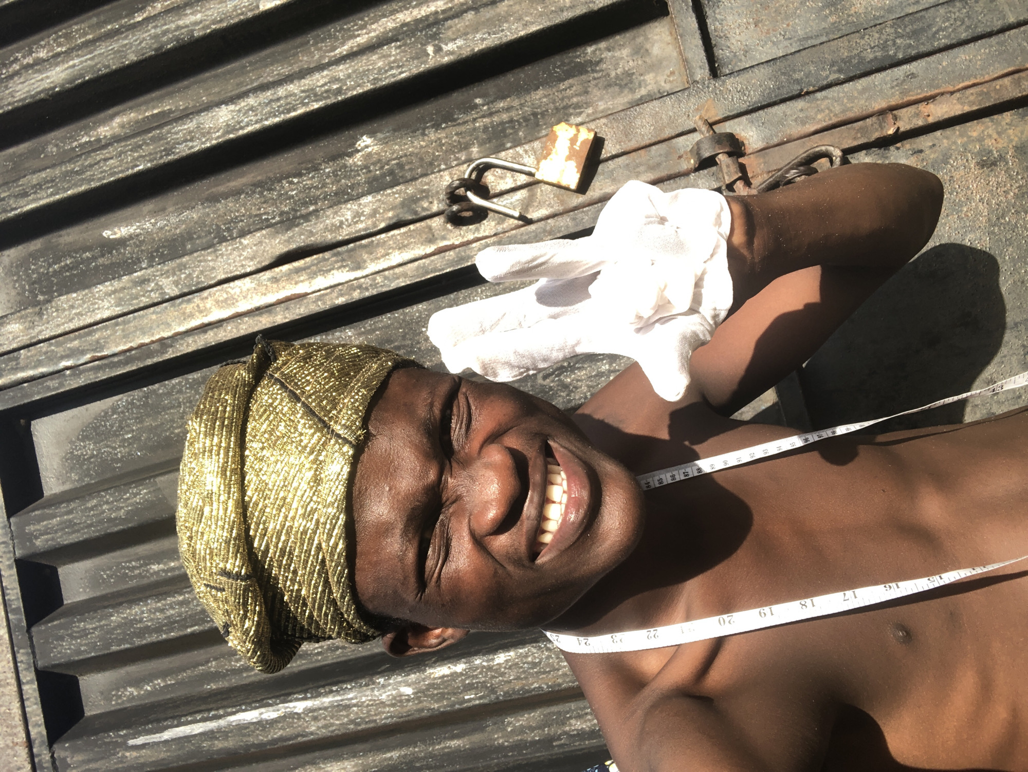 A young Black man with dark skin and a gold, textured headwrap is posing for a selfie in front of a corrugated metal surface. He is shirtless, and a white measuring tape is draped around his neck. He is wearing a white glove on his right hand and is holding it up in a peace sign. His eyes are squinted and he is smiling broadly, showing his teeth. A rusty padlock is visible on the metal surface to the right of the man. The scene appears to be outdoors, with strong sunlight casting shadows.