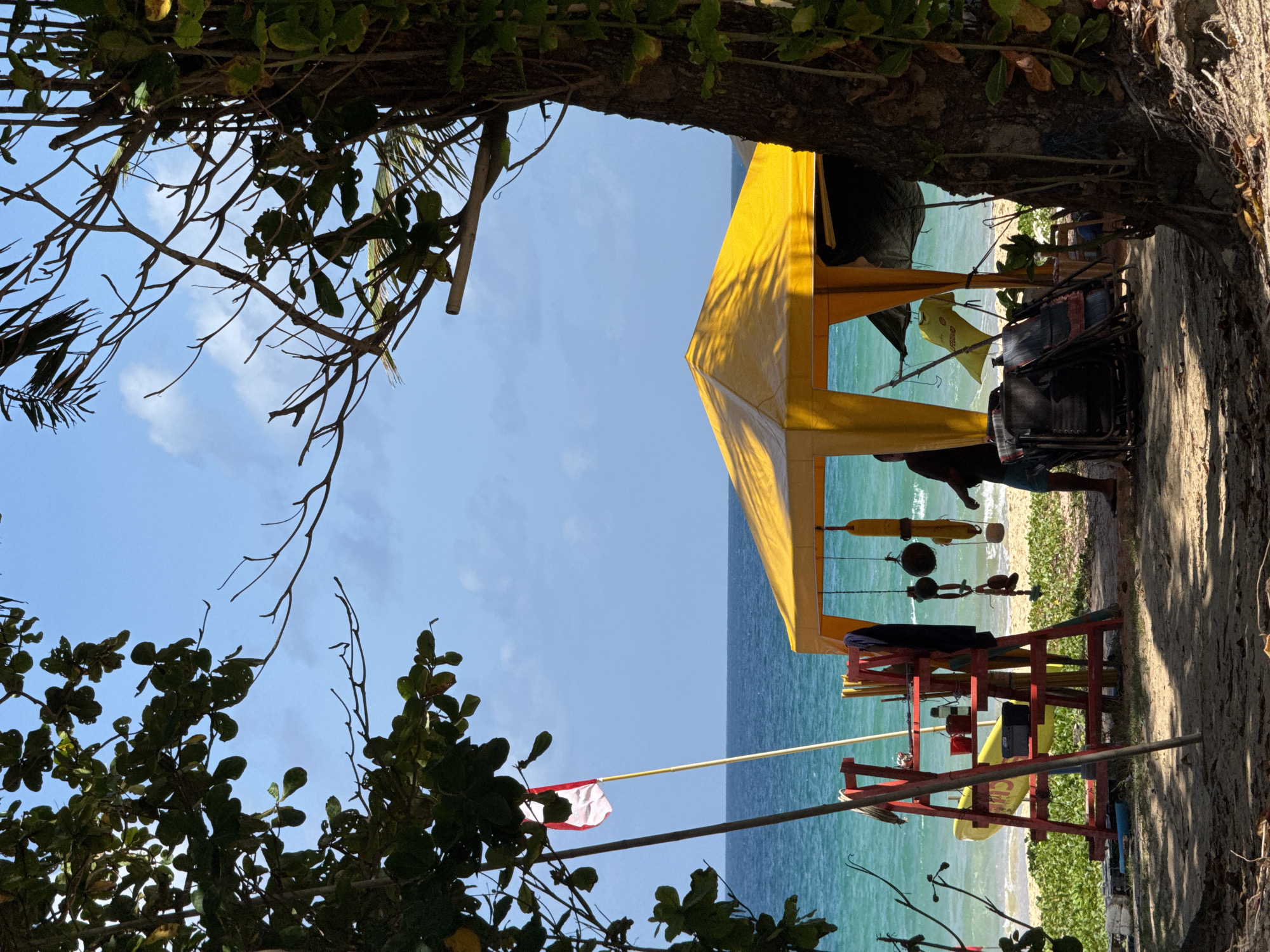 This image depicts a beachfront scene in Karon, Thailand, likely on a sunny day with a clear blue sky and scattered clouds. In the foreground, a sandy area is partially covered by the dappled shade of trees. To the left, a red lifeguard stand is visible, equipped with various items including what appears to be a yellow surfboard, red buckets, and a black toolbox. A red and white flag is planted on a pole next to the stand.
Dominating the mid-ground is a bright yellow canopy tent. Hanging from its structure are several objects, including what look like cooking pots and a yellow t-shirt. A person, silhouetted against the bright background, stands partially within the tent. Next to the tent, a stack of folded chairs is visible. The turquoise ocean stretches out in the background, with gentle waves lapping the shore. Lush green vegetation grows along the edge of the beach. The overall impression is one of a relaxed, tropical beach setting.