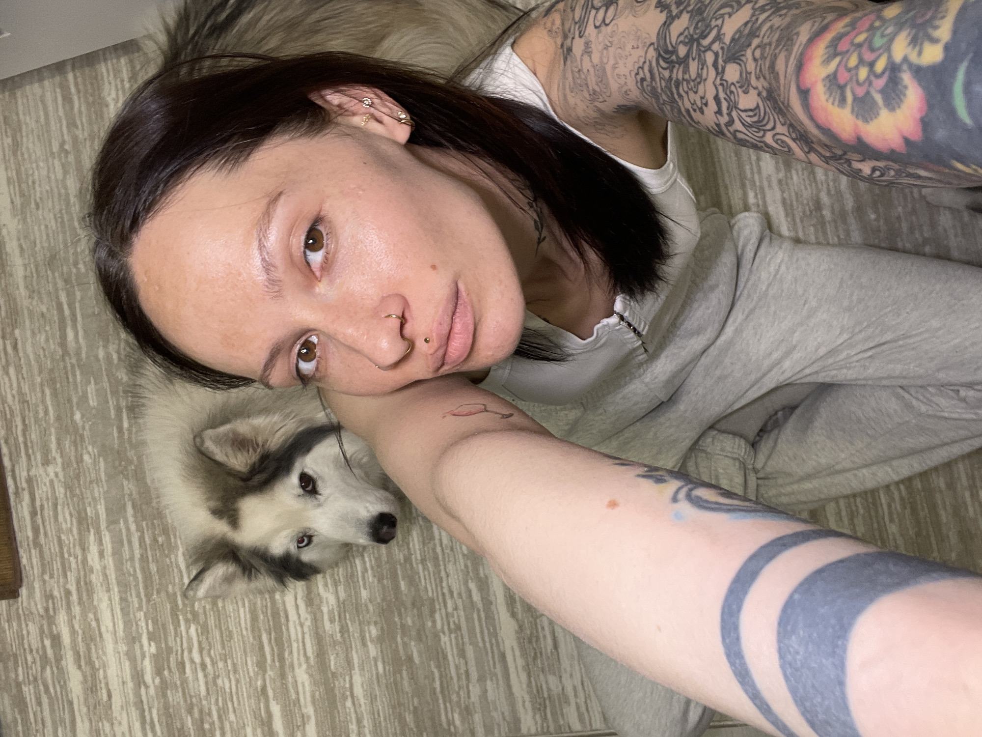 A high-angle, overhead shot depicts a young woman and a dog resting indoors on a light-colored, patterned floor. The woman, who has dark hair pulled back, brown eyes, and fair skin, is positioned on her side, with her head leaning on her right arm (viewer's left). She features multiple facial piercings, including a gold hoop in her left nostril and a small stud in her septum, as well as several earrings in her left ear. Both of her arms are extensively tattooed; the upper left arm (viewer's right) displays a vibrant, colorful design with yellow, orange, and red elements, while the lower right arm (viewer's left) shows a distinct black and grey abstract or geometric pattern. She is dressed in a light-colored tank top and light grey sweatpants.
Next to her, a fluffy dog, appearing to be a Husky or Malamute mix with distinctive white, grey, and black markings and dark, attentive eyes, gazes up towards the camera. The woman maintains a neutral, calm expression. The floor beneath them features a light-toned, wood-grain or laminate pattern. The image captures a relaxed, casual moment between the woman and her canine companion. There is no visible text.