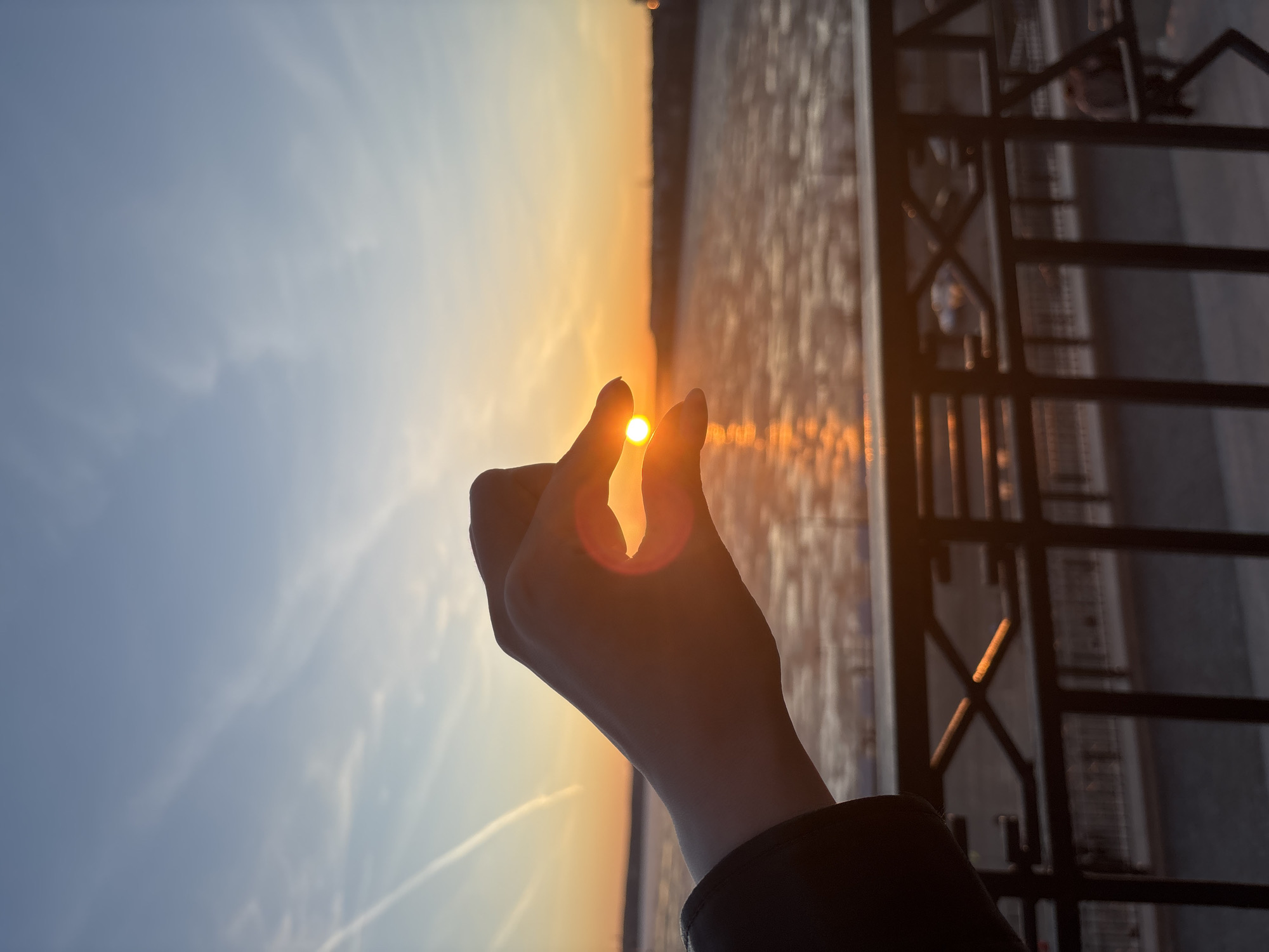 A vertical shot captures a playful moment during sunset in Izhevsk, Russia. The silhouette of a human hand dominates the center-right of the frame, with its index finger and thumb positioned to seemingly "pinch" or "hold" the bright, setting sun. A strong sun flare emanates from the sun, creating a warm, golden glow around the fingers and wrist.
The sky occupies the majority of the background, transitioning from a clear, light blue at the top and left to a vibrant orange and yellow where the sun is setting. Wispy clouds are visible, along with a distinct white contrail from an aircraft in the lower left of the sky.
To the right, a dark, vertical metal railing or fence is visible, partially silhouetted against the bright sky and the golden light reflecting off the background. The scene beyond the railing is blurred, suggesting a distant landscape, possibly urban features or a body of water, bathed in the warm light of the setting sun. The overall impression is one of serenity and creative interaction with nature at dusk. There is no visible text in the image.