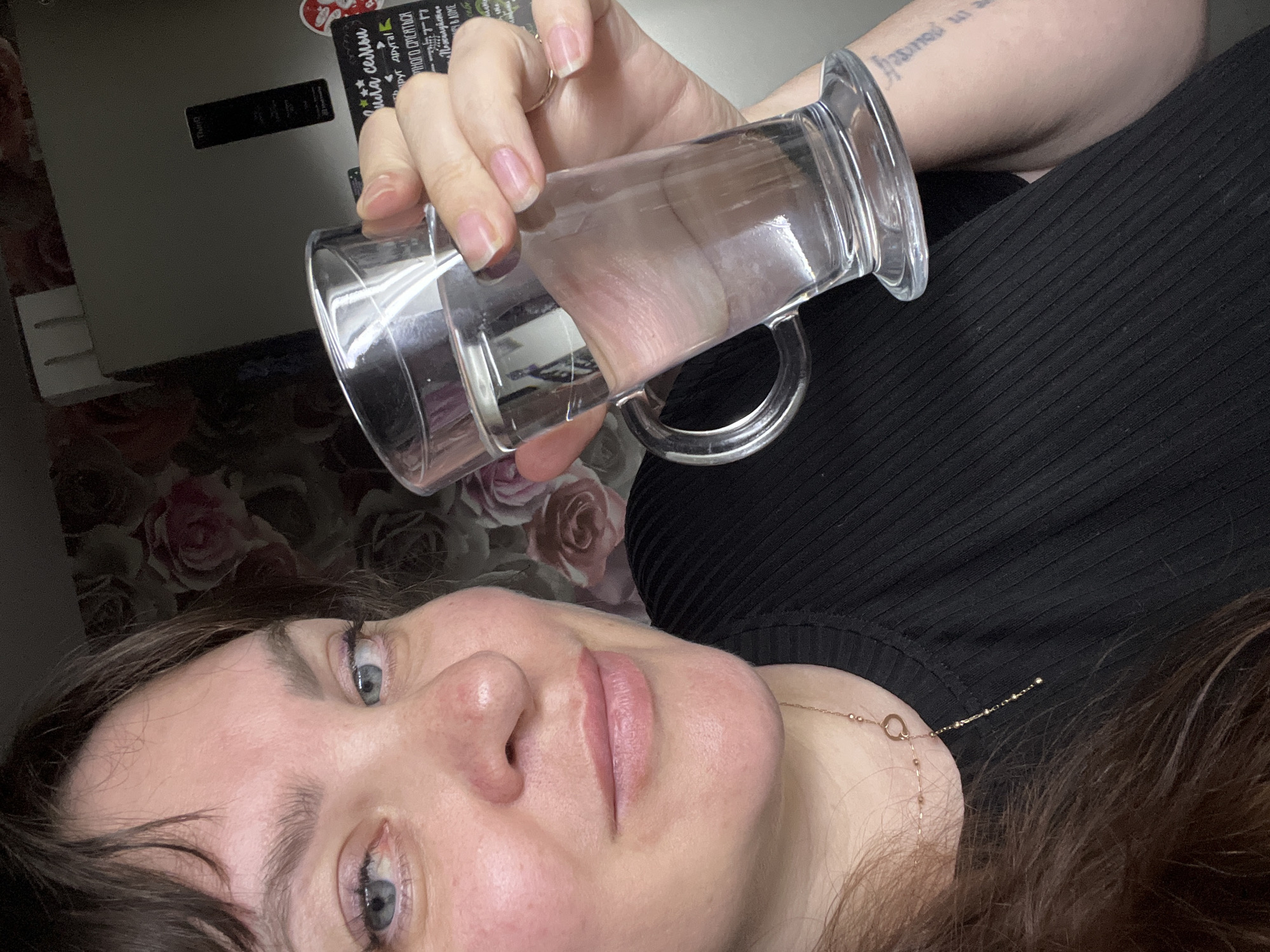 An adult female is depicted in a close-up, indoor shot, positioned in the foreground looking directly at the viewer with a slight smile. She has dark brown hair with bangs, blue eyes, and fair skin with some redness on her cheeks and around her nose. Her left eye shows slight redness on the lower eyelid. She is wearing a black ribbed top, a delicate gold-colored necklace, and a simple gold-colored ring on her right ring finger. Her nails are natural and unpolished. A partial text tattoo is visible on her left forearm/upper arm, with the phrase "in yourself" partially legible.
She is holding a clear glass mug, filled with clear liquid, in her right hand, which is positioned prominently in the center right of the frame. The glass has a handle and tapers upwards from a wider base.
The background shows a domestic interior, likely a kitchen or dining area, in the city of Noviy, Russia. Behind the woman, a wall is covered with floral wallpaper featuring large, soft-focus pink and white roses on a darker ground. To her right, a light gray/silver LG ThinQ refrigerator is visible. Attached to the refrigerator door are several items: a black board with white Cyrillic text, including phrases such as "семьи" (family), "Друг друга" (each other), "много смеяться" (laugh a lot), "Пожалуйста" (Please), and "в доме" (in the house/home). A red heart-shaped sticker is also affixed to the refrigerator door.