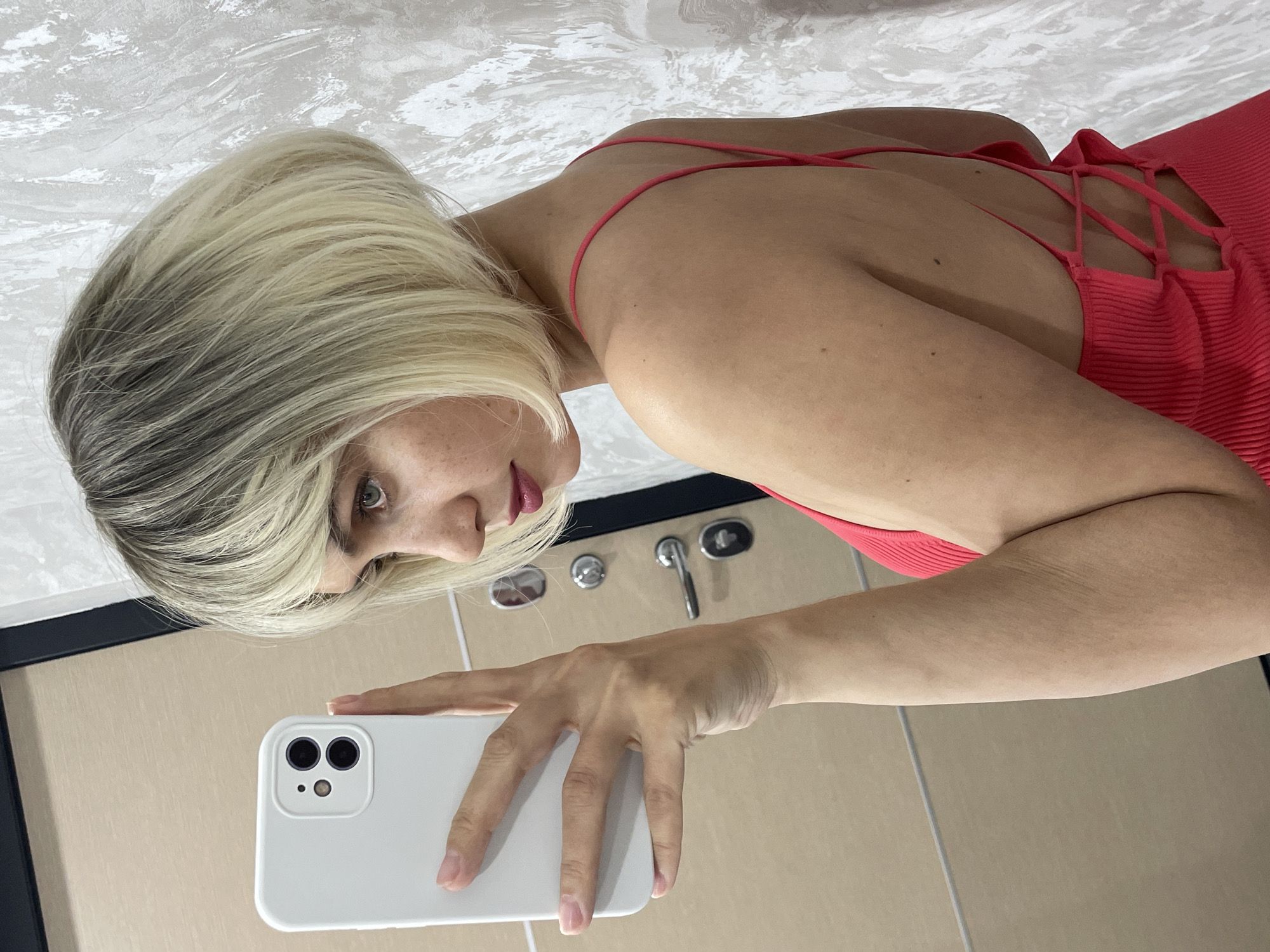 A young adult female takes a mirror selfie indoors, likely in a bathroom, in Izhevsk, Russia. She is light-skinned with blonde hair showing darker roots and visible freckles on her nose and cheek. Her lips are painted a rosy pink/red. She wears a form-fitting, ribbed coral or red dress or top, characterized by thin spaghetti straps and a criss-cross lace-up design on the back, visible from her leaning posture.
Her right hand holds a white smartphone, likely an iPhone with a dual-camera system, directed towards the mirror to capture her reflection. The reflected background includes a light-colored, possibly textured wall or ceiling with subtle patterns, and light brown/beige tiled surfaces on what appears to be the floor, with visible grout lines. Reflected bathroom fixtures, specifically a faucet and two control knobs, are also visible behind her hand. The woman's gaze is directed towards her reflection, with a neutral expression. The setting appears to be lit by artificial indoor lighting. No visible text is present in the image.