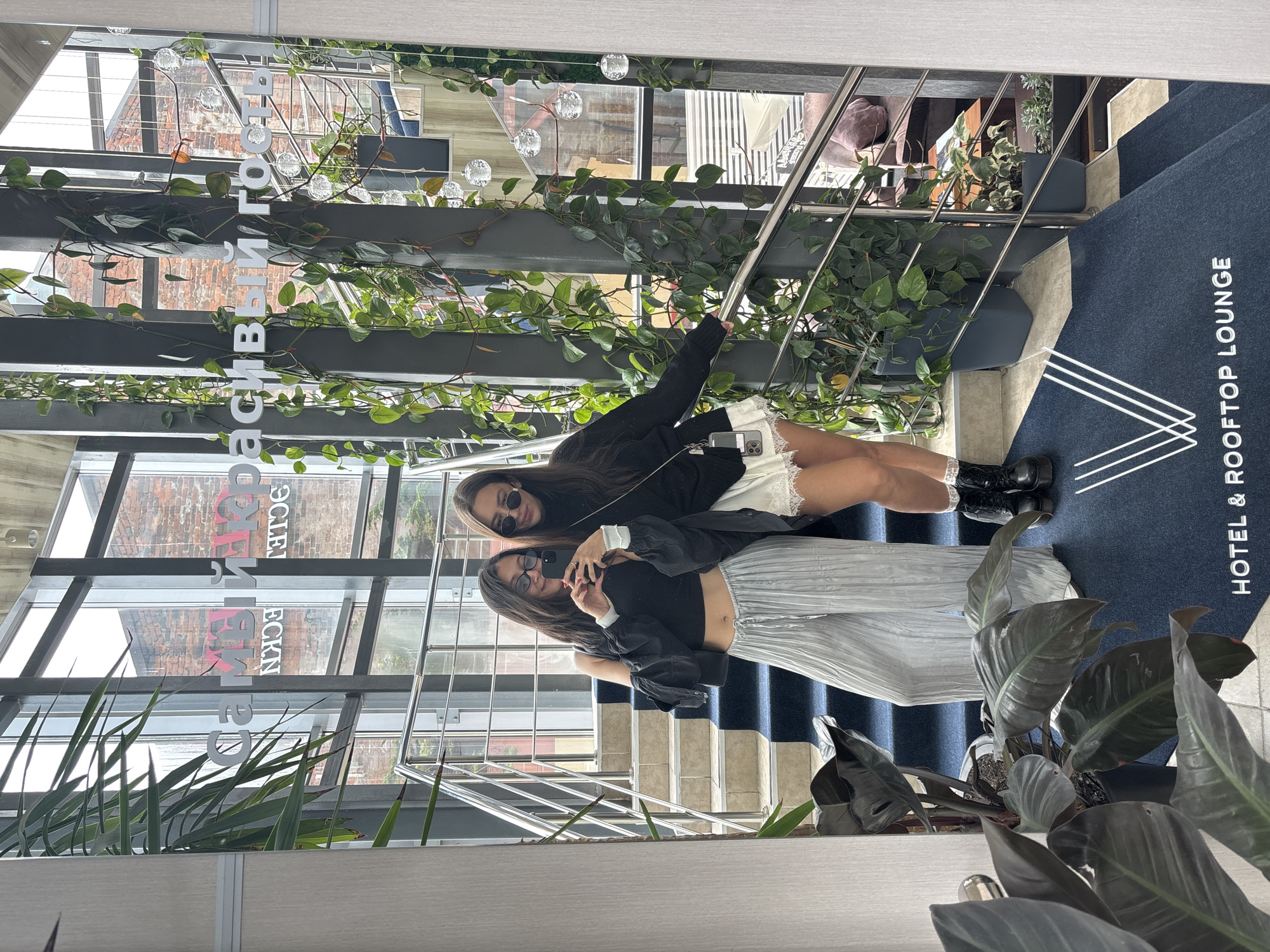 Two women pose for a photo on a staircase, with "HOTEL & ROOFTOP LOUNGE" printed on the floor near the stairs. One woman wears a black top, silver pants, and sunglasses while holding a phone. The other wears a black jacket, a white skirt, black boots, and sunglasses. Plants are along the side of the stairs, along with a reflective ceiling with text that reads "DECK" and "КРАСИВЫЙ ГОРОД". The location is Vladivostok, Russia.