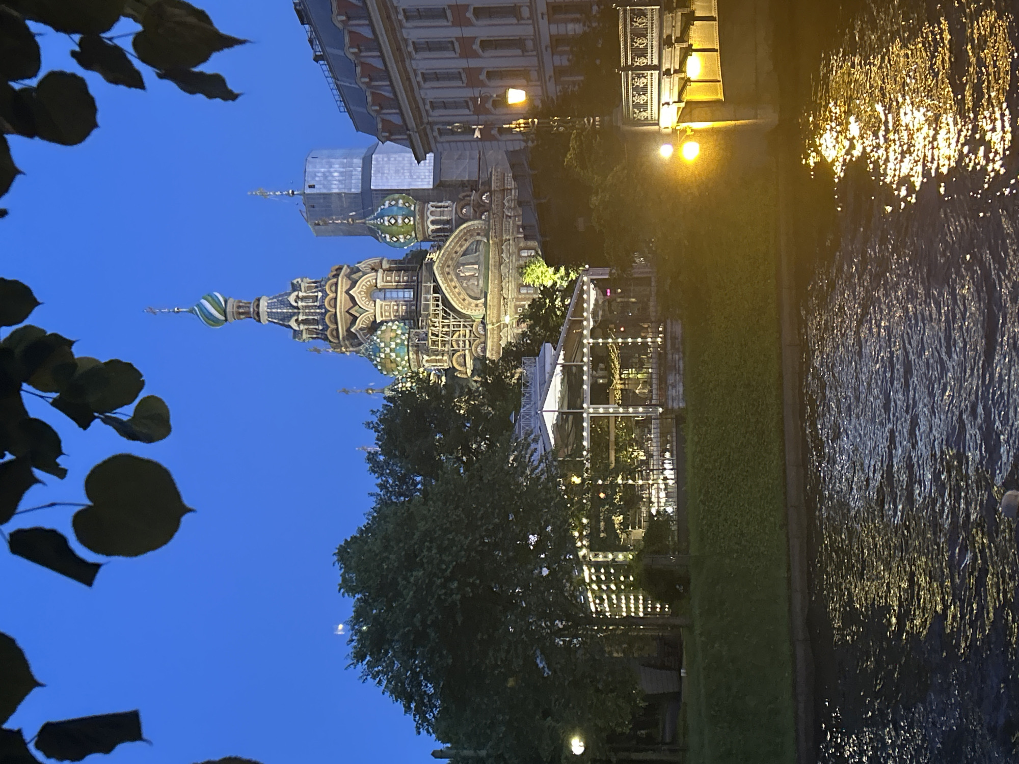 The image depicts a twilight scene along a waterway with a distinctive onion-domed cathedral in the background. The cathedral, identified as the Church of the Savior on Spilled Blood, is illuminated by artificial lights, highlighting its ornate architecture and colorful domes. In the foreground, the dark water reflects the yellow glow of streetlights and lights from a structure that appears to be an open-air cafe or seating area. The cafe is adorned with numerous strings of small, white lights. Trees with dense foliage are visible on the grassy embankment between the waterway and the buildings. A bridge with decorative railings is also partially visible on the right side of the image, with more streetlights casting warm illumination. Dark leaves hang from branches in the upper part of the frame, framing the sky, which is a deep, clear blue. There are no people clearly visible in the image.