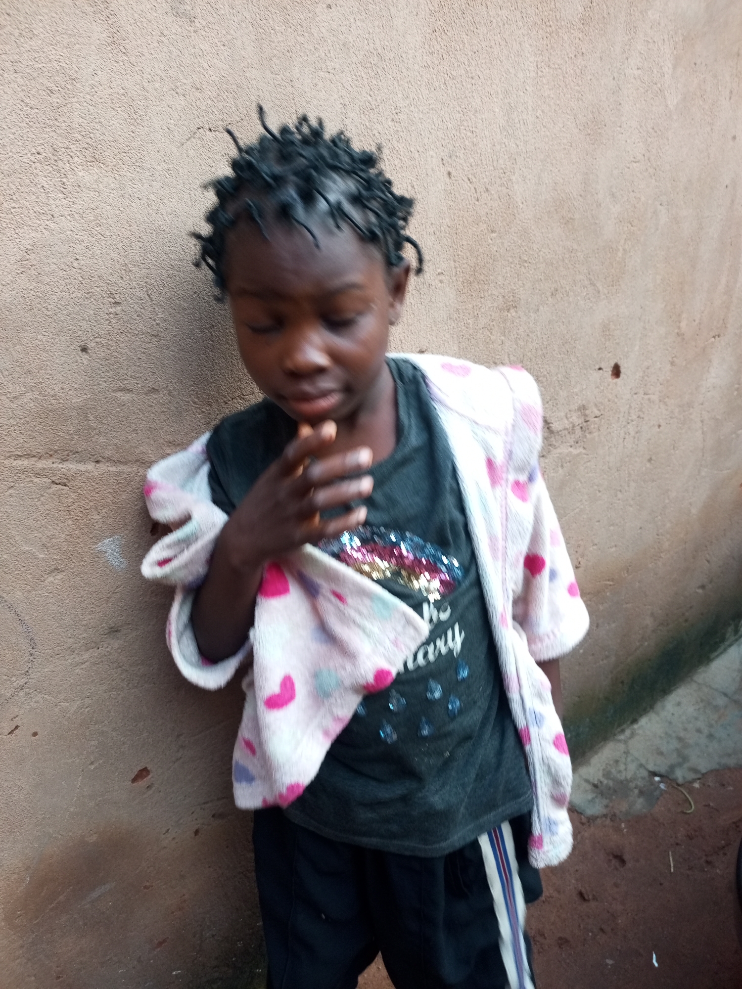 The image shows a young Black child, likely a girl, standing against a textured beige wall. She is wearing a dark gray t-shirt with some sparkly embellishments on the chest and black shorts with a white and red stripe down the side of each leg. A light pink robe with small hearts in pink and purple is draped over her shoulders. Her hair is styled in short, dark dreadlocks. The child is looking down, with her right hand raised towards her mouth. The wall behind her appears to be outdoors, as evidenced by the textured surface and a hint of ground and shadow to the right. The overall impression is of a casual, everyday scene, possibly in a residential area of Tempure, Nigeria.