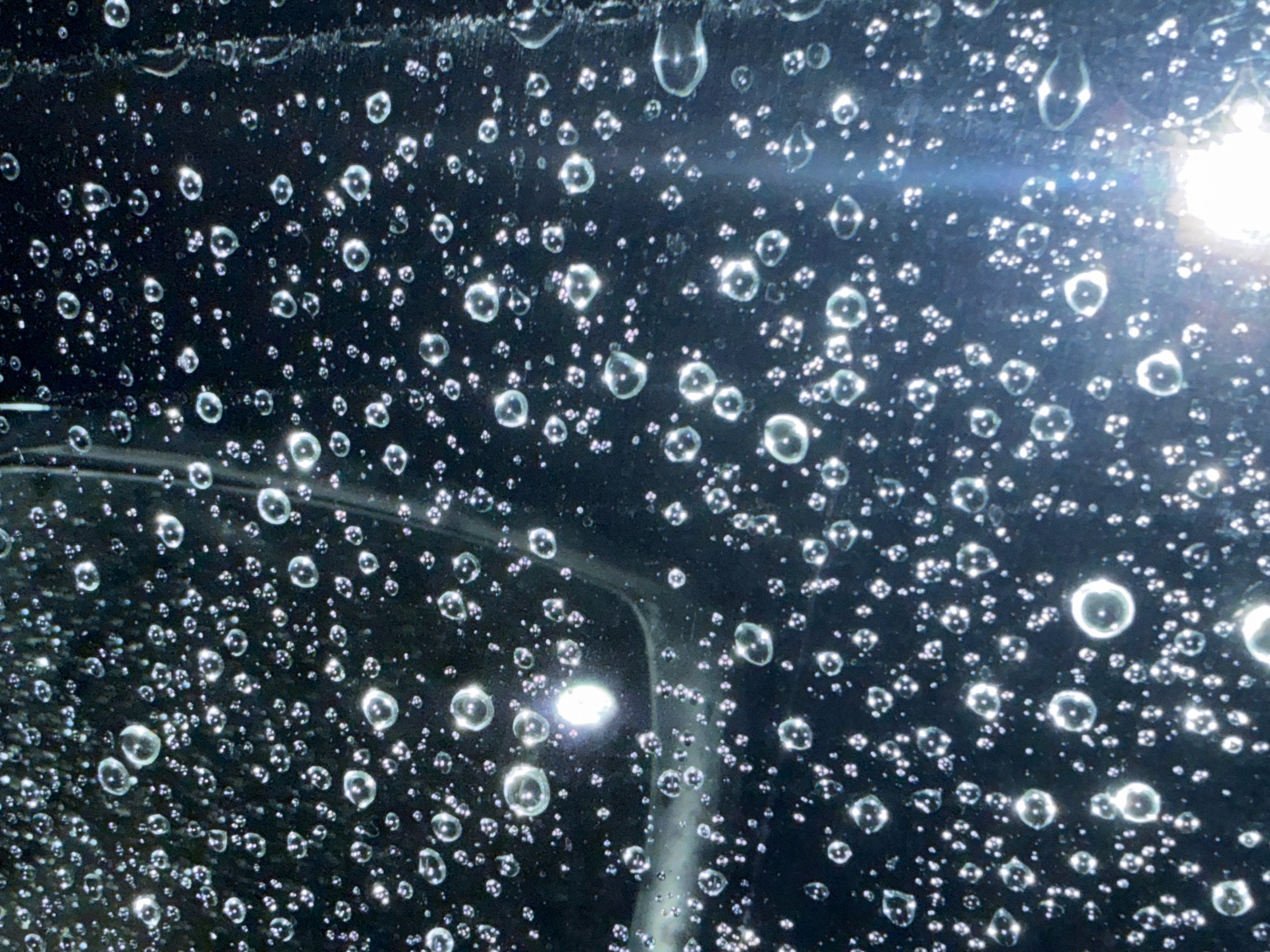 The image shows a close-up of water droplets on a dark, possibly reflective surface. The droplets vary in size, from tiny specks to larger, more rounded shapes, and they are illuminated by a bright light source, causing reflections and highlights. The surface appears to be textured, with some streaks or lines visible beneath the droplets, suggesting it might be glass or a similar material. The scene is abstract and focuses on the texture and light play of the water. There are no people or identifiable city elements visible. The location in Тосор, Kyrgyzstan, is not visually represented in the image itself.