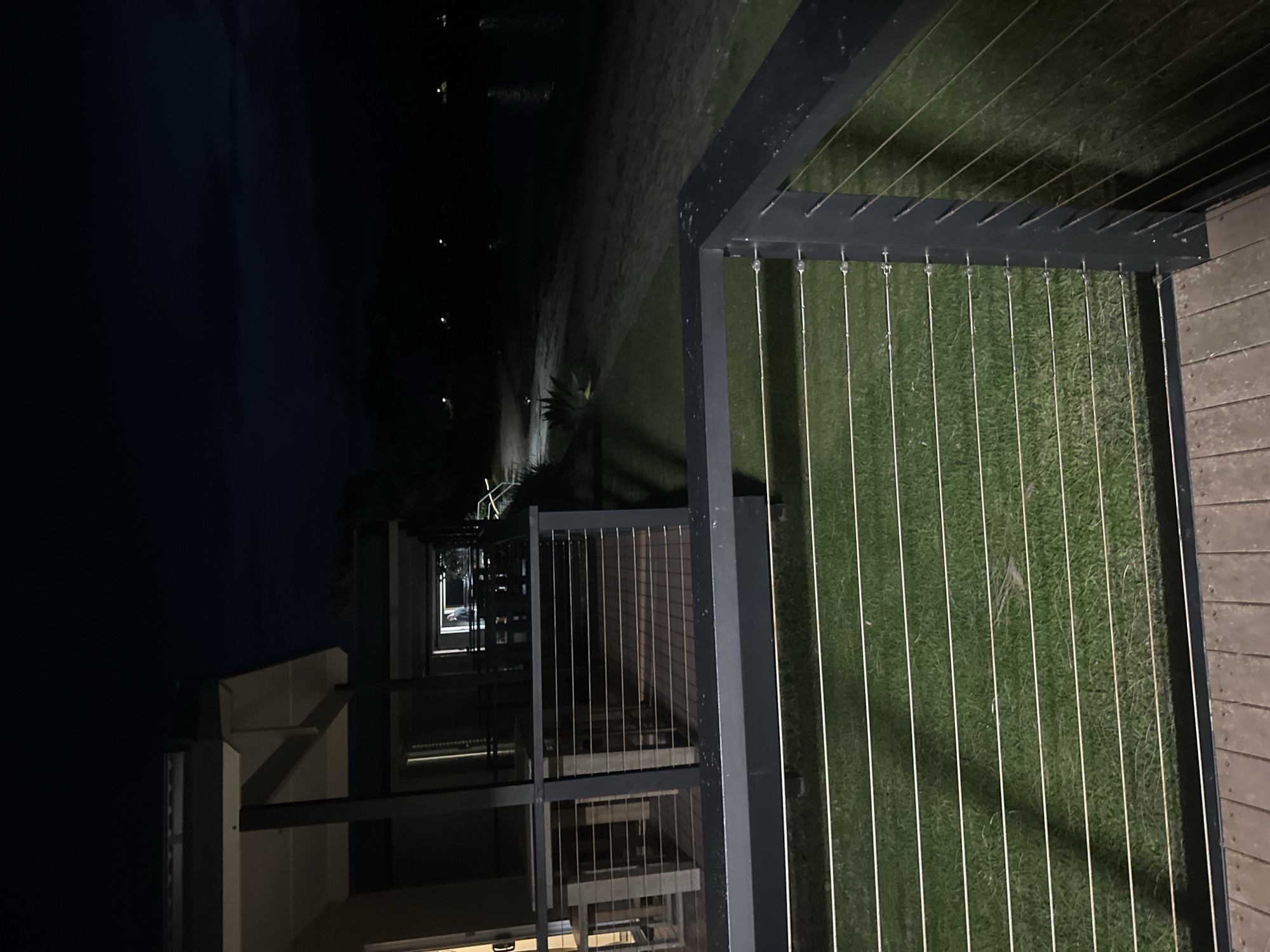 This is a night time, low-light, vertical, medium-shot photograph taken from a wooden deck. In the foreground, a dark grey metal railing with thin horizontal wires runs across the bottom of the frame. Beyond the railing, a neatly mowed green lawn is visible. A wooden deck is also visible in the foreground, made of wide wooden planks.
In the middle ground, another deck extends to the left, with a dark grey railing and horizontal wires. Tables and chairs are visible on this deck. Further in the background, a building with white and dark grey trim is visible. Lights are on inside, illuminating the interior. A person is visible standing in a doorway, silhouetted against the interior light.
In the background, a sandy area stretches horizontally across the frame, with some sparse vegetation. Beyond the sand is a body of dark water, possibly a river or ocean. Distant lights are visible along the shoreline, suggesting a populated area. The sky above is a deep, dark blue, indicating nighttime. The overall scene suggests a resort or residential property located near a waterfront in Nambucca Heads, Australia, possibly a holiday park or motel.