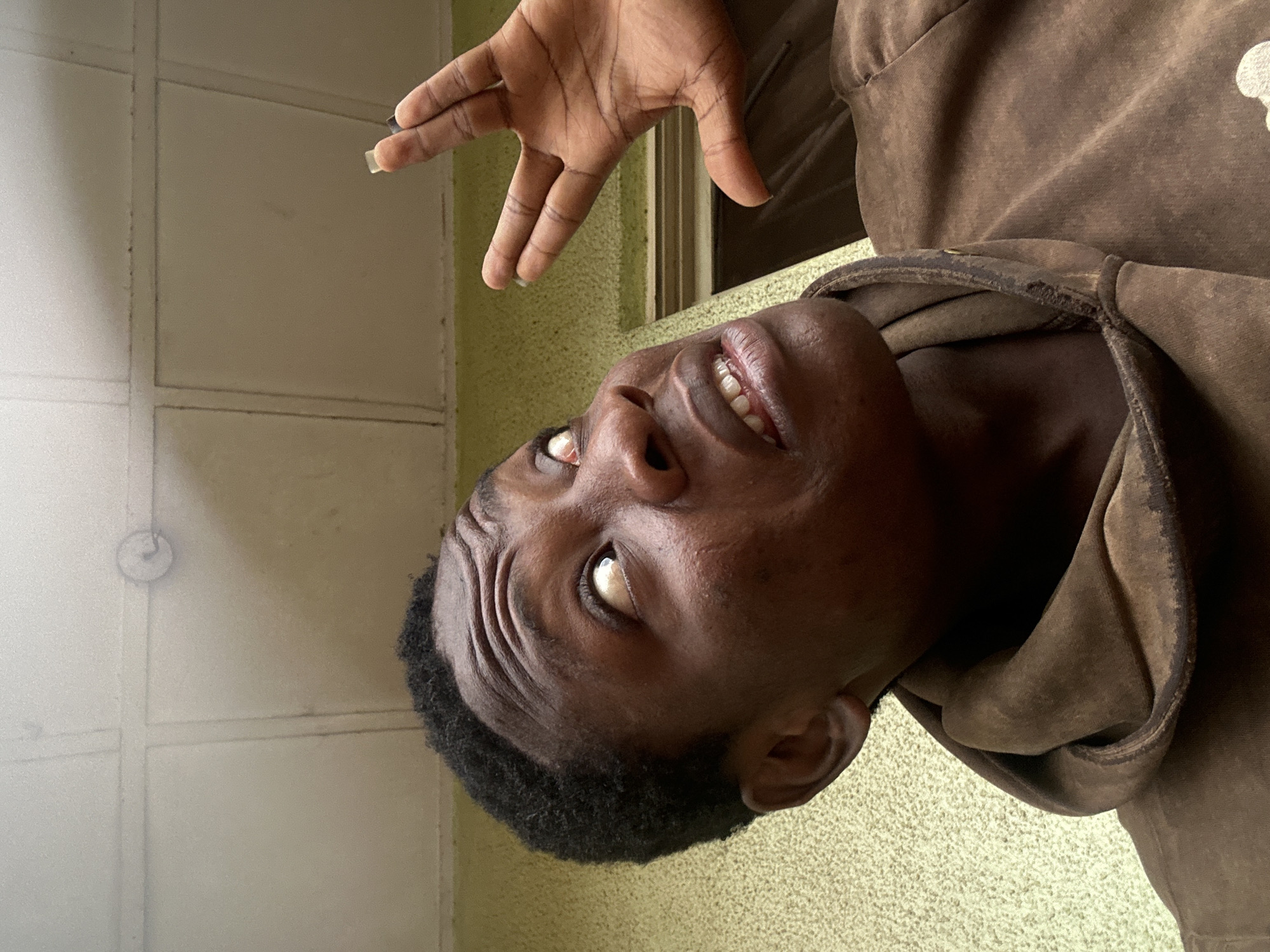 A dark-skinned person wearing a brown hooded sweatshirt is looking upwards with their mouth slightly open. Their hand is raised above their head, fingers spread. The background includes a light green wall and a white panelled wall. The location is Unknown, Unknown.