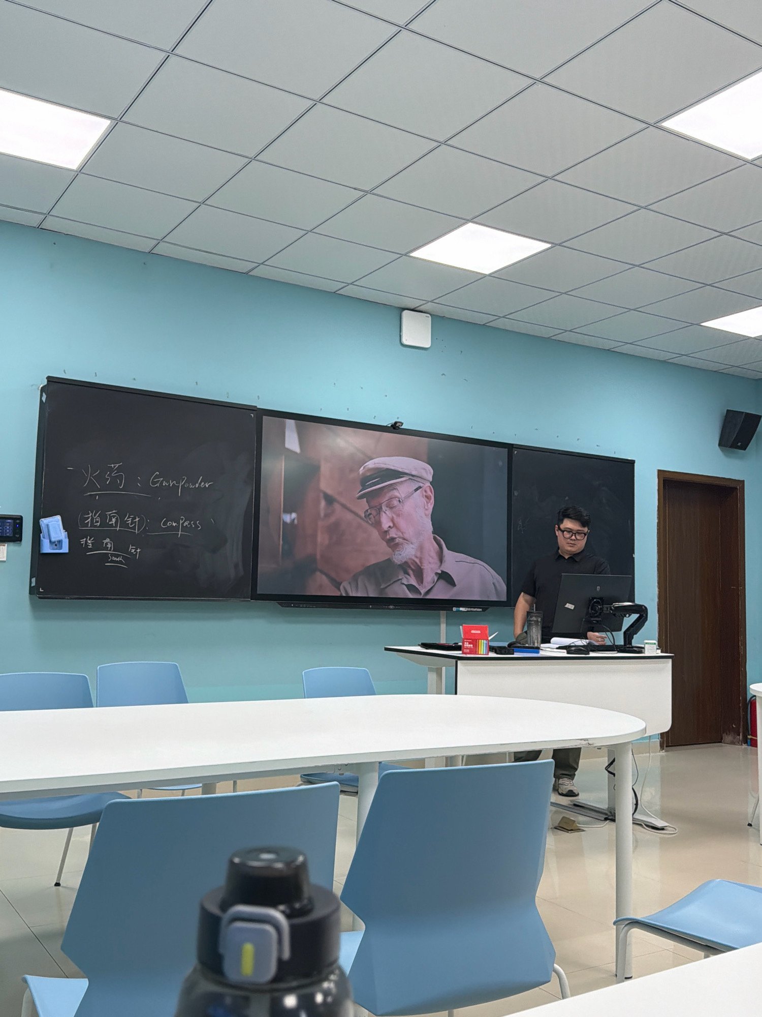 The image depicts a classroom setting in Jiangjin, China, featuring a teacher standing at a podium in front of a large screen displaying a video of an elderly man. A blackboard is mounted to the left of the screen, with some handwritten Chinese characters and English words. The classroom is equipped with rows of light blue chairs and white tables. Fluorescent lighting panels are visible on the ceiling. The room's walls are painted a light blue color. The teacher, a young man wearing glasses and a black polo shirt, is engaged with a computer monitor on the podium. The overall environment suggests an educational or presentation scenario.