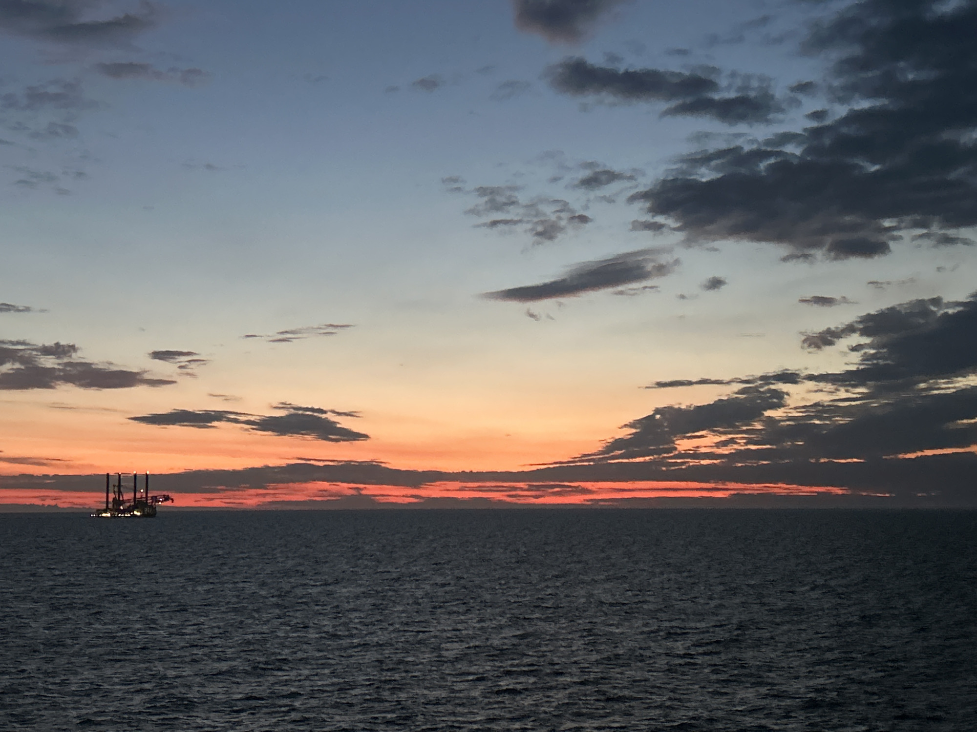 This image depicts an offshore scene at dusk or dawn, located in Unknown, Unknown. The sky transitions from a light blue at the top, through a band of muted grey-blue with scattered dark grey clouds, to a vibrant horizontal strip of orange and red near the horizon. Prominent dark grey clouds, some forming distinct horizontal layers, are concentrated on the right side of the frame and above the horizon. The lower half of the image is dominated by a vast expanse of dark, rippled ocean water, exhibiting subtle surface texture from small waves. On the left side, an industrial offshore platform is situated against the illuminated horizon. The platform appears as a dark silhouette, but several bright lights are visible on its structure, illuminating parts of its multiple vertical and horizontal components. No individuals or specific actions are discernible. The environment is open water under a dramatic sky.
