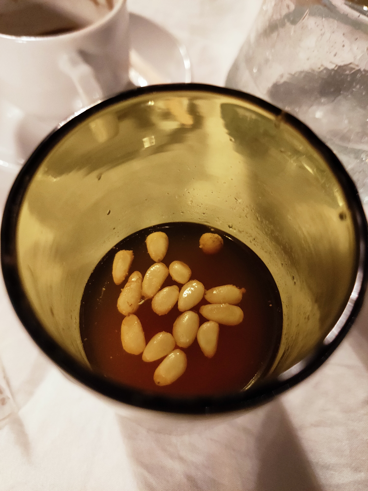 In Cebalat, Tunisia, a close-up, high-angle shot focuses on a clear glass containing a dark liquid and several pine nuts. The glass, with a dark rim, is positioned on a white, textured surface, likely a tablecloth. To the left of the glass, a white ceramic coffee cup and saucer are partially visible. To the right, a frosted glass containing ice and water is also in view. The lighting appears to be artificial, casting shadows and creating reflections on the glass surfaces. The scene suggests a casual setting, possibly a cafe or restaurant.