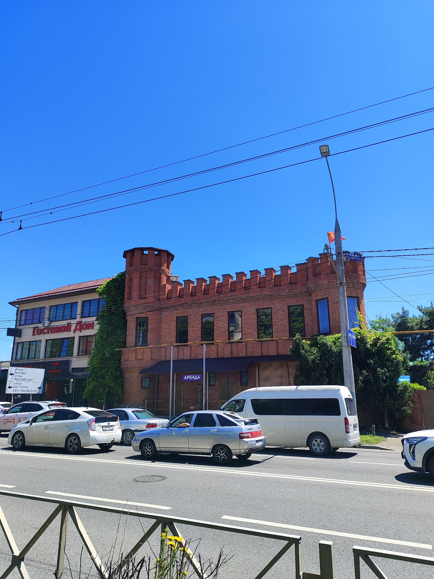 This image captures a vibrant street scene in Krasnodar, Russia, under a clear, bright blue sky on a sunny day, likely midday or early afternoon, given the distinct shadows.
In the midground, two prominent buildings line the street. To the left, a multi-story building with a red-tiled roof features a large sign in Cyrillic that reads "Гостевой Дом" (Guest House). Green ivy partially covers its facade. Below this, smaller Cyrillic text indicating "КУХНЯ" (Kitchen) and "АВТОШКОЛА" (Driving School) is partially visible. Adjacent to it on the right is a distinctive red-brick building designed with a castle-like aesthetic, including a round turret and a crenellated parapet along its roofline. This building also has ivy growing on its side, and a sign on its lower level reads "АРЕНДА" (Rent) in Cyrillic.
The foreground features a multi-lane asphalt road with clear double white lines. A metal barrier or fence runs along the bottom edge of the frame, separating the street from the viewer's position. Several cars are visible on the road: a white Toyota Prius, a silver sedan (likely a Lada), and a silver coupe (possibly a Toyota Soarer/Lexus SC). Further right, a white minivan (resembling a Toyota Hiace) and the front of a white SUV are also present. No people are distinctly visible.
Overhead, multiple power lines crisscross the sky, and a tall street light pole with a modern LED fixture stands on the right side of the image. To the far left, a white street sign with a left-pointing arrow lists several street names in Cyrillic: "УЛИЦА ЖЛОБЫ" (Zloby Street), "УЛИЦА РОССИЙСКАЯ" (Rossiyskaya Street), and "ВОСТОЧНО-КРУГЛИКОВСКАЯ" (Vostochno-Kruglikovskaya Street).