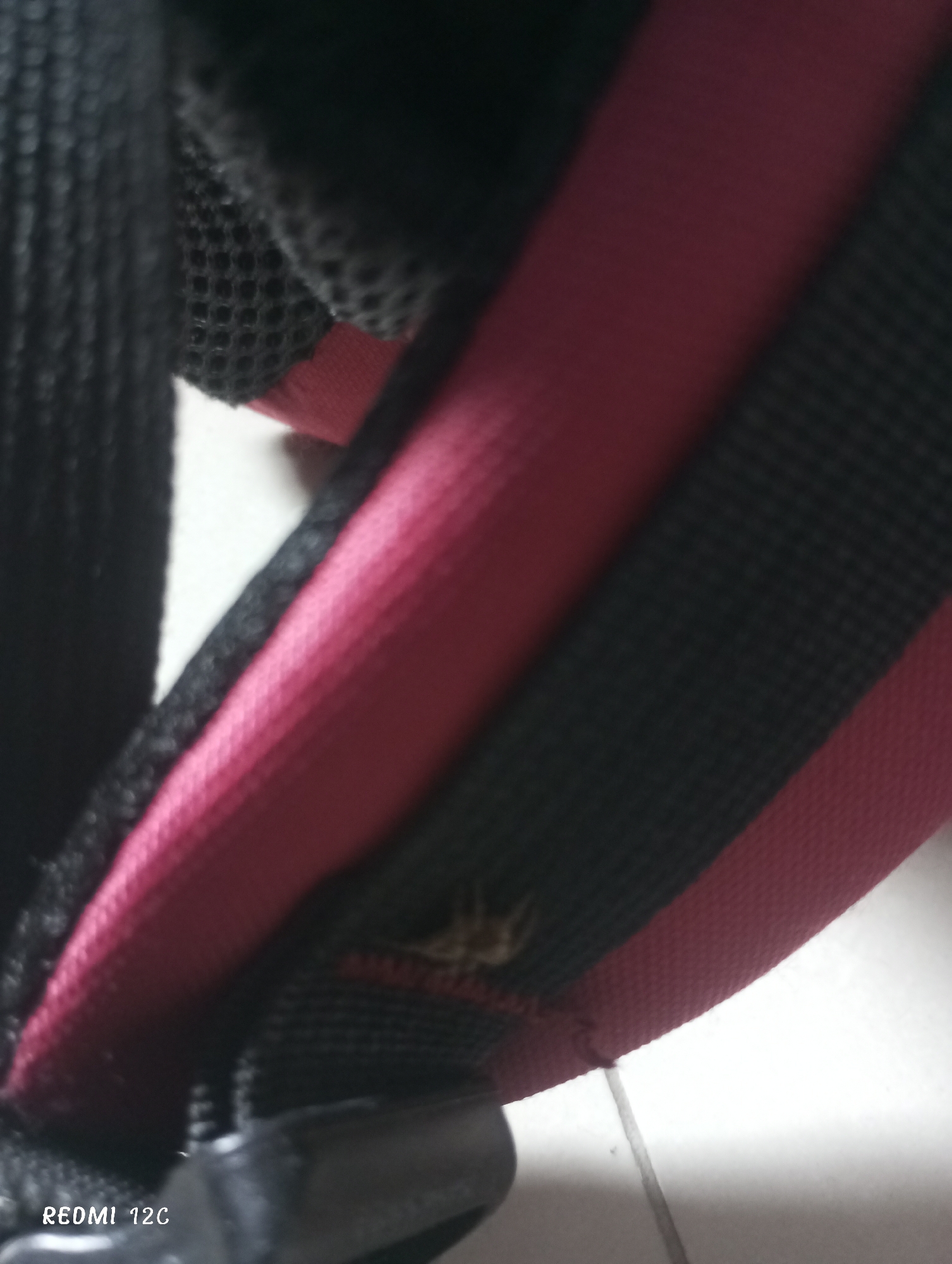 The image shows a close-up of a black and red backpack strap. The strap has a black mesh portion and a solid red portion. A portion of the strap is connected to a plastic buckle. The image was captured using a Redmi 12C camera. The location of the image is unknown.