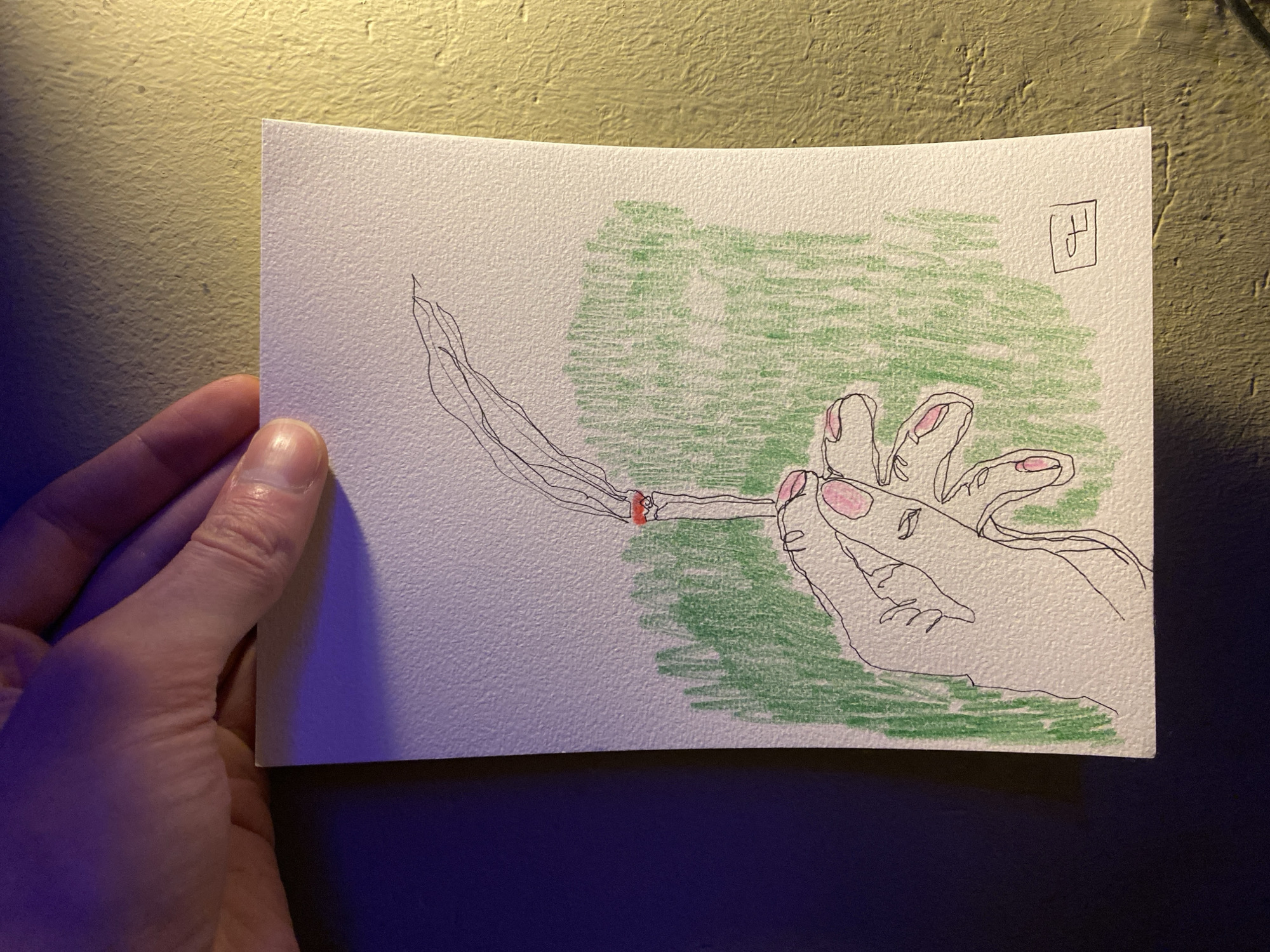 A close-up photograph captures a hand presenting a small, artistic drawing on textured white paper. The drawing features a stylized, continuous line art depiction of a human hand, appearing to be a left hand, holding a lit, hand-rolled cigarette. The cigarette has a bright reddish ember at its tip and emits prominent, wispy smoke trails extending upwards and to the left. The drawn hand shows subtle pink coloring on the fingernails.
The background within the drawing is partially filled with loose, horizontal scribbles of green crayon or colored pencil, creating an abstract, textured effect. In the top right corner of the drawing, a small square contains a stylized character, possibly an "H," serving as an artist's signature or monogram.
The actual hand holding the drawing is visible in the lower left and bottom edges of the photograph. The scene is illuminated by artificial light, with a warm, golden-yellow light source from the upper left casting light on a rough, stucco-textured wall behind the drawing. A cooler, purplish-blue light source appears to be coming from the lower right, influencing the colors on the real hand and the surface below the drawing. The paper's texture is clearly visible, suggesting heavy cardstock or art paper.