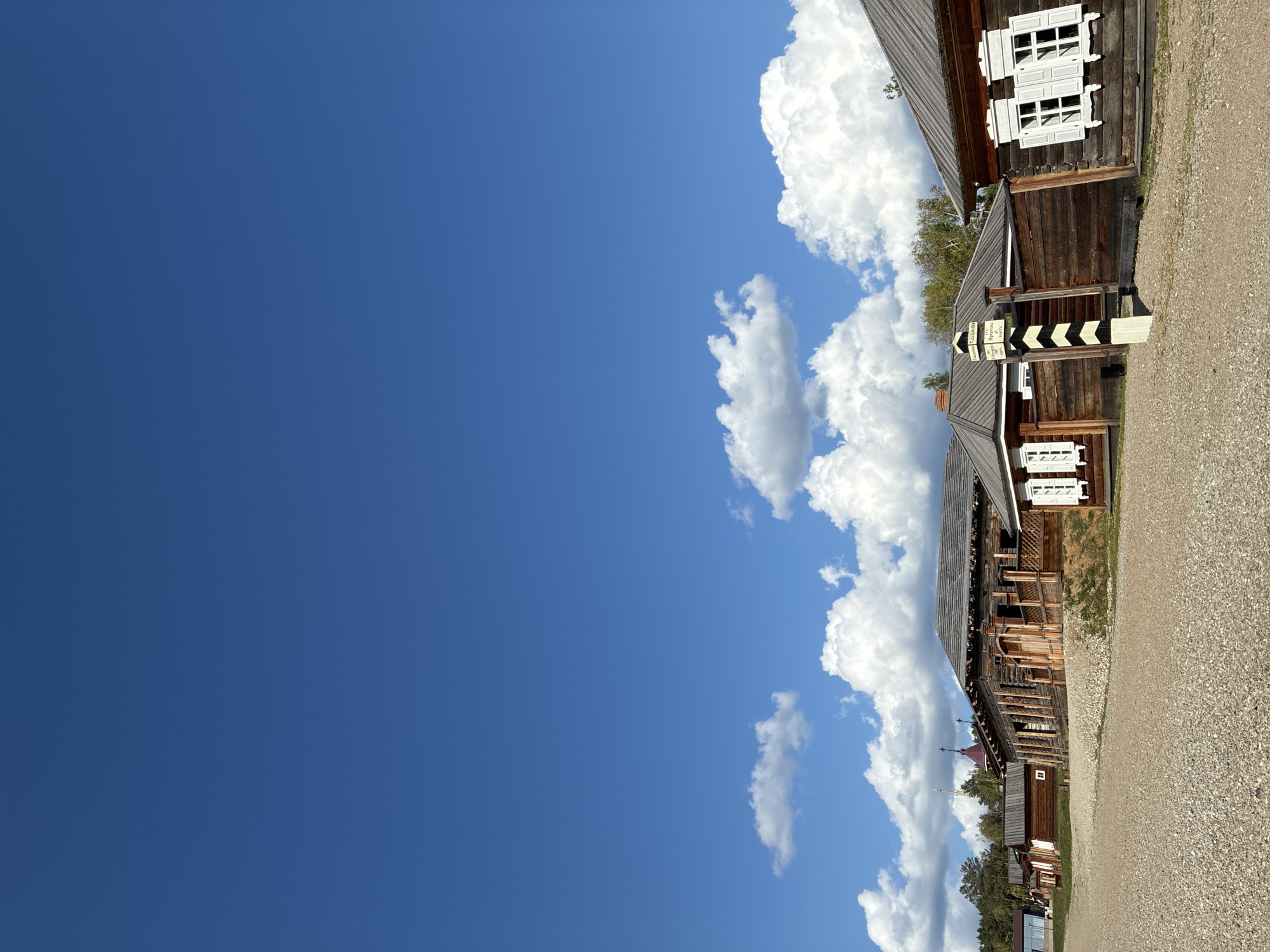The image depicts a cluster of traditional wooden buildings in the open-air museum of Taltsy, located near Irkutsk, Russia. The structures feature dark wooden walls and roofs, with white-trimmed windows, some of which have closed shutters. A gravel pathway runs in front of the buildings. In the foreground, a black and white striped pole with inscriptions is visible. The sky is a clear blue with scattered white clouds. The overall scene conveys a historical and rustic atmosphere.