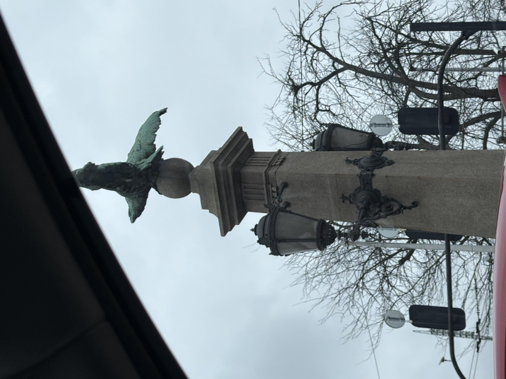 A low-angle shot shows a statue atop a stone column, possibly of an eagle, situated at the intersection of Tsar Osvoboditel Boulevard and Georgi S. Rakovski Street in Sofia, Bulgaria. The statue and column feature ornate detailing, including mounted lamps on the column and decorative elements at its base. The background includes bare tree branches against an overcast sky. A portion of a dark structure or vehicle interior is visible in the lower left, partially obstructing the view, while a red vertical object appears on the right side of the column. The surrounding environment seems urban.