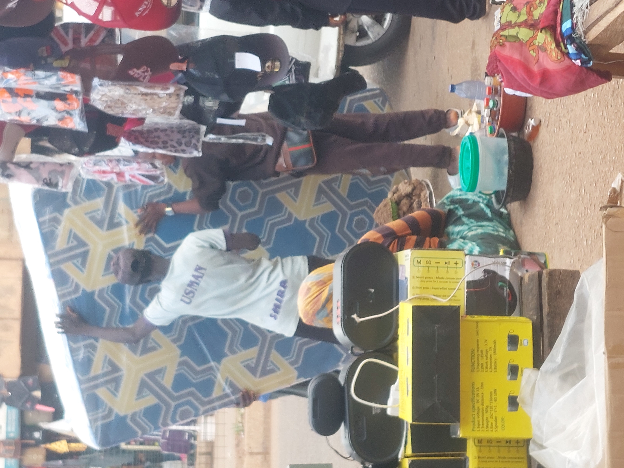 The scene depicts an active outdoor street or market environment in the city of Unknown, Unknown. Three individuals are collaboratively handling a large, rectangular object, likely a mattress or foam board, approximately 2 meters in length and 1.5 meters in height. This object features a blue background adorned with a repeating pattern of yellow geometric cuboid shapes.
The central figure, viewed from behind, wears a light-colored t-shirt with "USMAN" printed prominently in purple capital letters across the upper back, and "SMIRA" inscribed below. He wears a dark baseball cap and grips the top edge of the patterned board with his raised left hand. To his right, another male figure, also seen from behind, is dressed in a dark brown long-sleeved shirt and matching pants, with a brown and green striped fanny pack positioned on his left hip. He supports the board with his left hand, which displays a wristwatch. A third person's hand is partially visible on the far left, also assisting with the board's movement.
In the foreground, a seated individual, with their upper body obscured, wears a multi-colored headscarf (yellow, blue, and orange tones) and a striped garment on their visible arm. This individual is positioned amidst a collection of merchandise displayed at ground level. This merchandise includes several black portable speakers resting on yellow cardboard boxes. One box explicitly details "Product specifications" and "FUNCTION," listing features such as "Input voltage: DC 5V 1A," "Transmission distance: 10m," "Weight: 80g," "Battery: 1800mAh," and available colors as "Green" and "Blue." White charging cables are connected to some of the speakers. Nearby, a black bowl contains dark, granular items, while a green-lidded container sits within another black bowl. A brown bowl holds multiple small, stoppered bottles and several light-colored, peeled food items. A clear plastic water bottle stands beside these items. A red and green patterned fabric piece is folded on a wooden structure to the far right.
On the right side of the image, numerous items, including various caps and small bags, hang from an unseen display fixture. Identifiable items include a cap featuring a Union Jack flag design, a leopard-print accessory, and a red cap with the letters "ANL" partially visible. The rear portion of a white vehicle, exhibiting a silver wheel rim, is visible on the far right. The ground surface is a light brownish-gray, suggestive of an unpaved area.