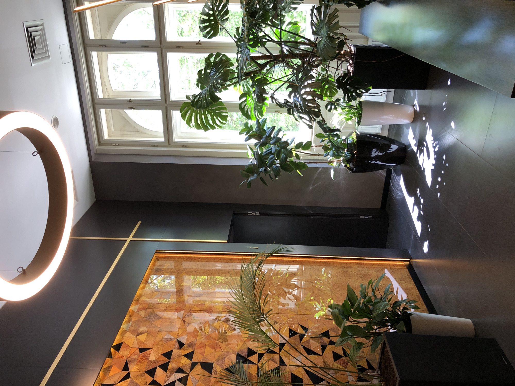 The image depicts an interior scene, possibly a lobby or waiting area, featuring modern design elements and lush greenery. A prominent circular light fixture with a warm glow hangs from the ceiling. The wall to the left is adorned with a striking geometric mosaic in warm tones of orange and brown, illuminated by vertical orange lighting. Several potted plants, including a large Monstera deliciosa, are strategically placed throughout the space, some in black planters and one in a white cylindrical pot. Sunlight streams in through a large, multi-paned window with arched upper sections, casting dappled shadows on the dark tiled floor. A dark door is visible to the right of the mosaic wall. The scene appears to be an upscale commercial or hospitality establishment, possibly in Lviv, Ukraine, given the context provided. There are no people or animals visible in the image.