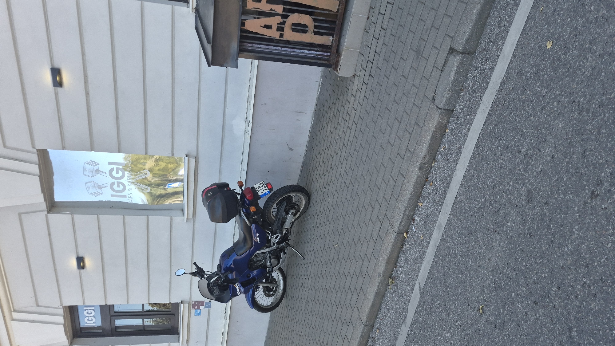 A blue Honda motorcycle is parked on a gray brick sidewalk in front of a white building with two large windows. The left window has "IGGI" written on it in white letters. The right window has a reflective surface with "IGGI BARS & KARBONADES" written on it in white letters, along with an image of two wrenches. The motorcycle has a black storage box on the back and a license plate reading "TD 7462". To the right of the motorcycle, a dark brown structure with vertical slats and the letters "AP" is visible. The building has horizontal lines and a modern outdoor light fixture. The street in front of the sidewalk is asphalt with a white line. The scene appears to be in a city, likely Daugavpils, Latvia, given the context. There are no people or animals visible in the image.
