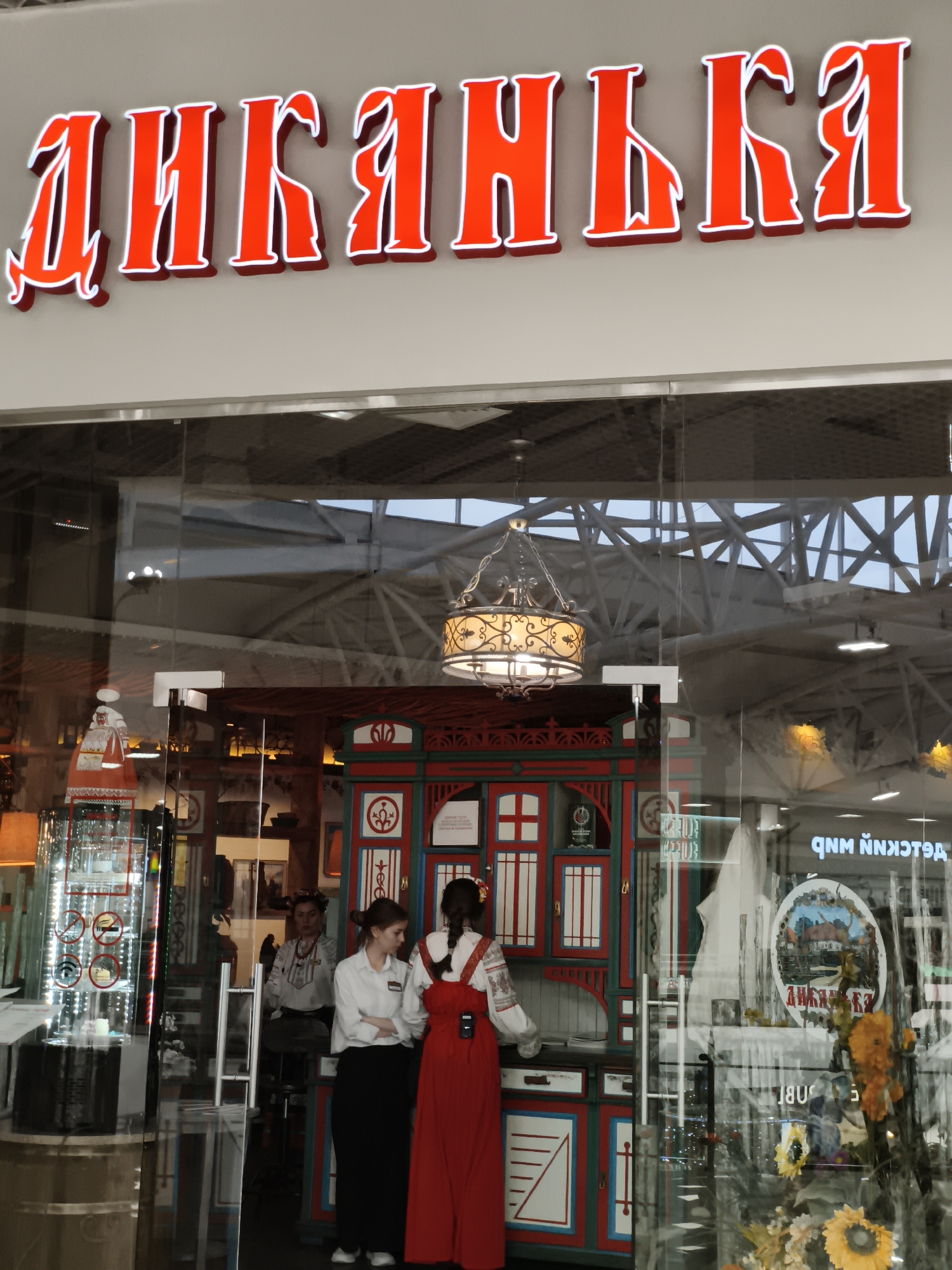 The image captures the entrance to a restaurant named "Диканька" in the city of Krasnodar, Russia. The restaurant's name is displayed in large red Cyrillic letters above the glass doorway. Inside the restaurant, two women are visible. One is wearing a white blouse and a long red dress with suspenders, while the other is wearing a white blouse and black pants. A third person sits on a stool in the background. The restaurant is decorated with a colorful, traditional cabinet, a decorative chandelier, and other decorative elements reflecting a Ukrainian theme. Through the glass, reflections of the surrounding indoor shopping center environment are visible.