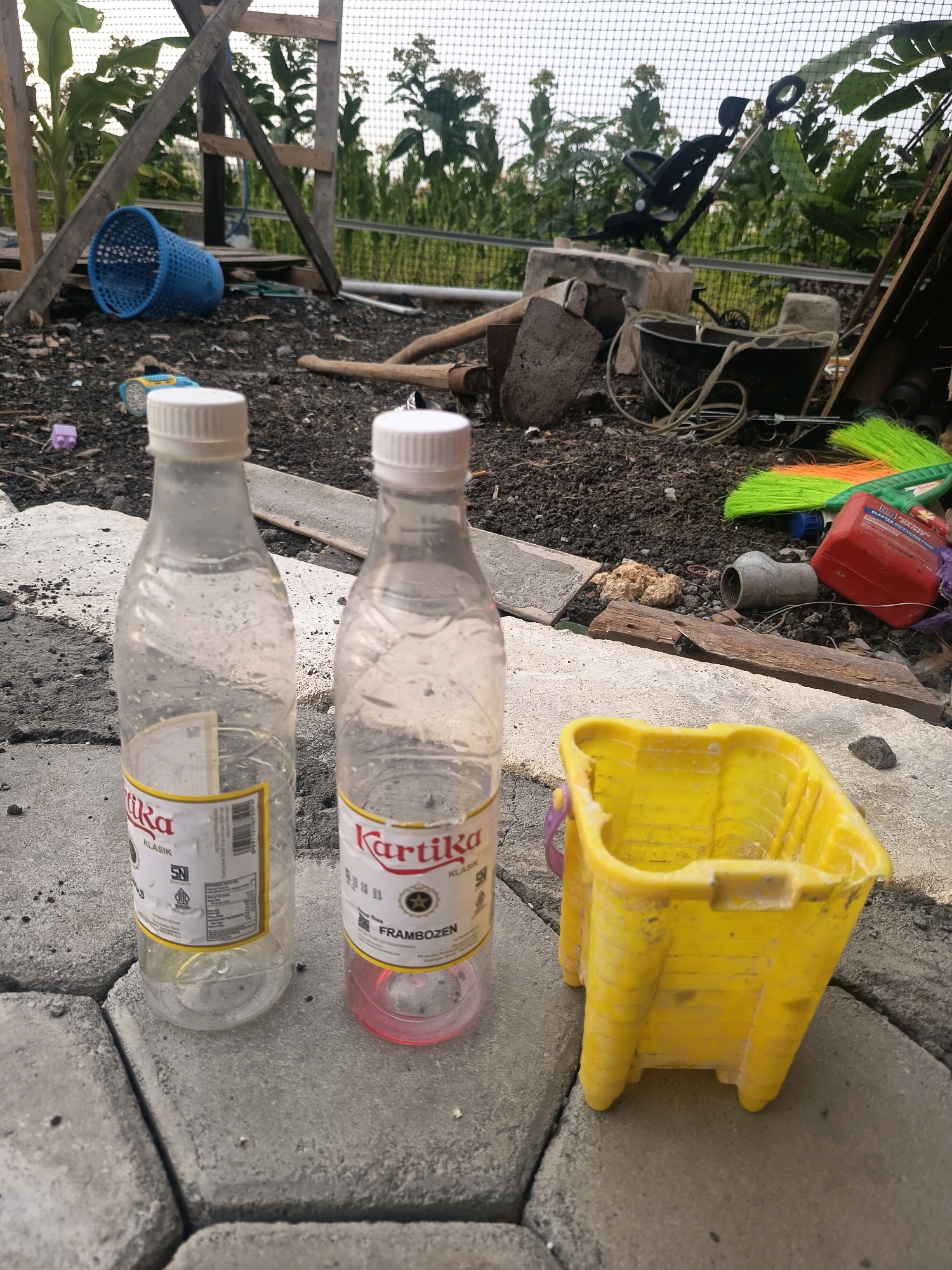 The image depicts a cluttered outdoor area, possibly a backyard or a yard in a rural or semi-rural setting, in Sidorejo, Indonesia. Two plastic water bottles, one mostly empty and the other containing a small amount of pink liquid, stand on a paved surface made of grey interlocking bricks. Next to the bottles is a bright yellow plastic bucket with purple handles. In the background, there is a wooden structure, a blue plastic basket, gardening tools including a shovel and a hoe, a black bucket, and a broom with green and orange bristles. Beyond these objects, vegetation is visible, including banana plants. A white net covers the upper part of the scene. The ground is covered with dark soil and debris. The overall impression is one of disarray and accumulated items in an outdoor space.