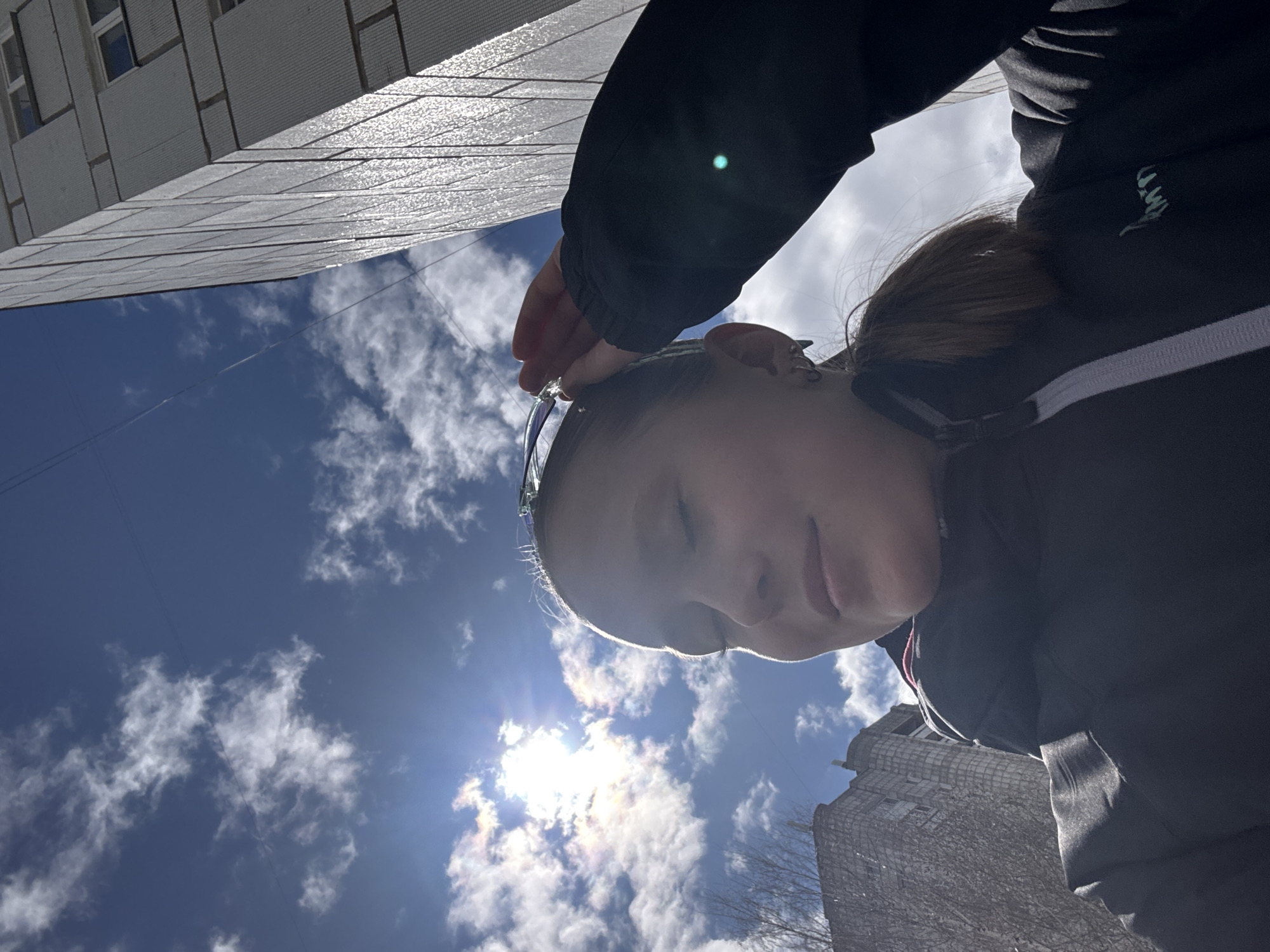 A low-angle shot features a person with sunglasses resting on their forehead, set against a backdrop of a partly cloudy, bright sky and buildings. The person is wearing a dark jacket with white stripes. The buildings exhibit a distinctive, possibly Soviet-era architectural style, which suggests the location may be the city of Chaikovsky, Russia. The time of day is daytime.
