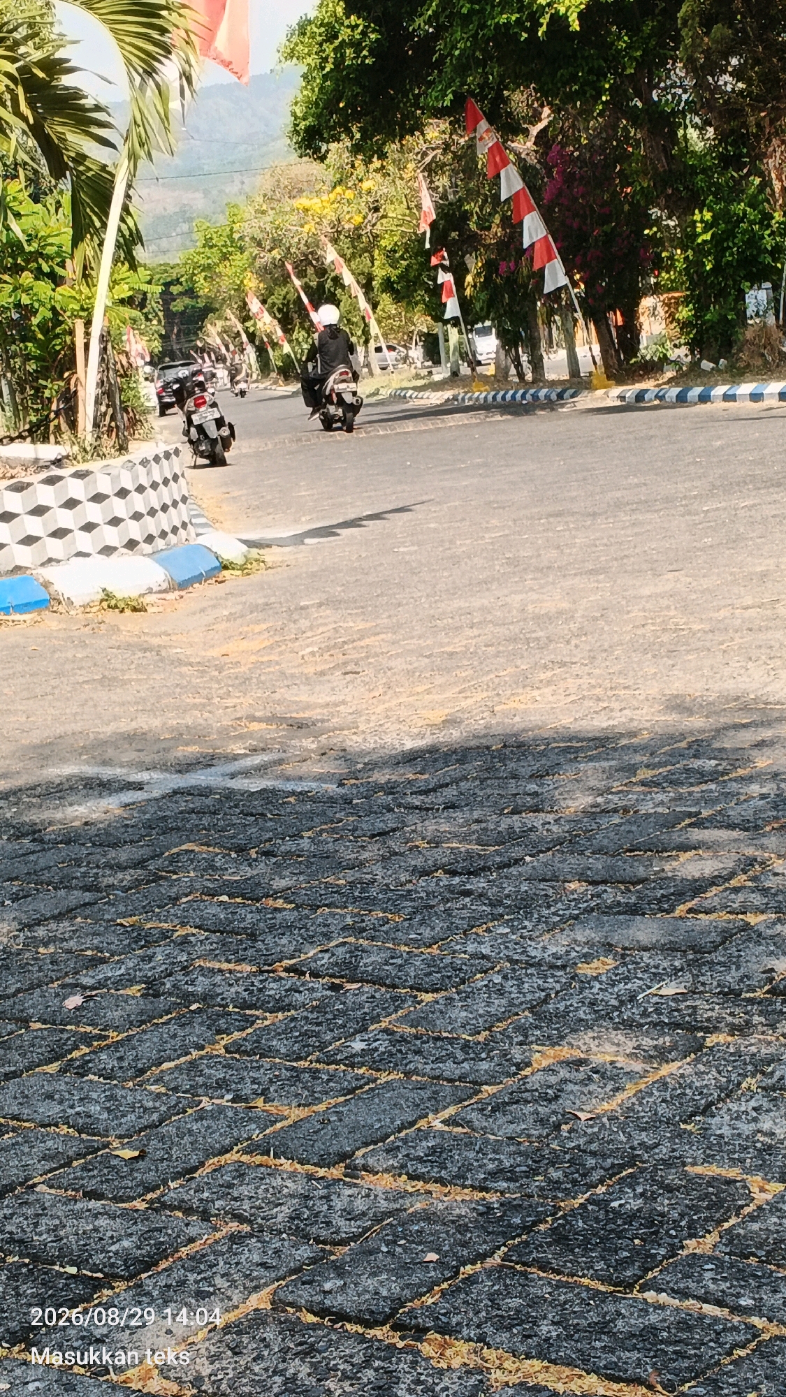 The image depicts a street scene in Mulyorejo, Indonesia, with a road paved with interlocking stones in the foreground, overlaid with scattered yellow petals. In the middle ground, a paved road has several motorcycles with riders, one of whom is wearing a white helmet and black attire, traveling in the same direction. The road is lined with a decorative concrete barrier on the left and a white and blue striped curb on the right. Red and white flags are prominently displayed on poles along the right side of the road, interspersed with trees. The background shows lush green foliage and distant hills under a clear sky. The overall impression is of a sunny day in a semi-urban or suburban area with a festive atmosphere due to the flags.