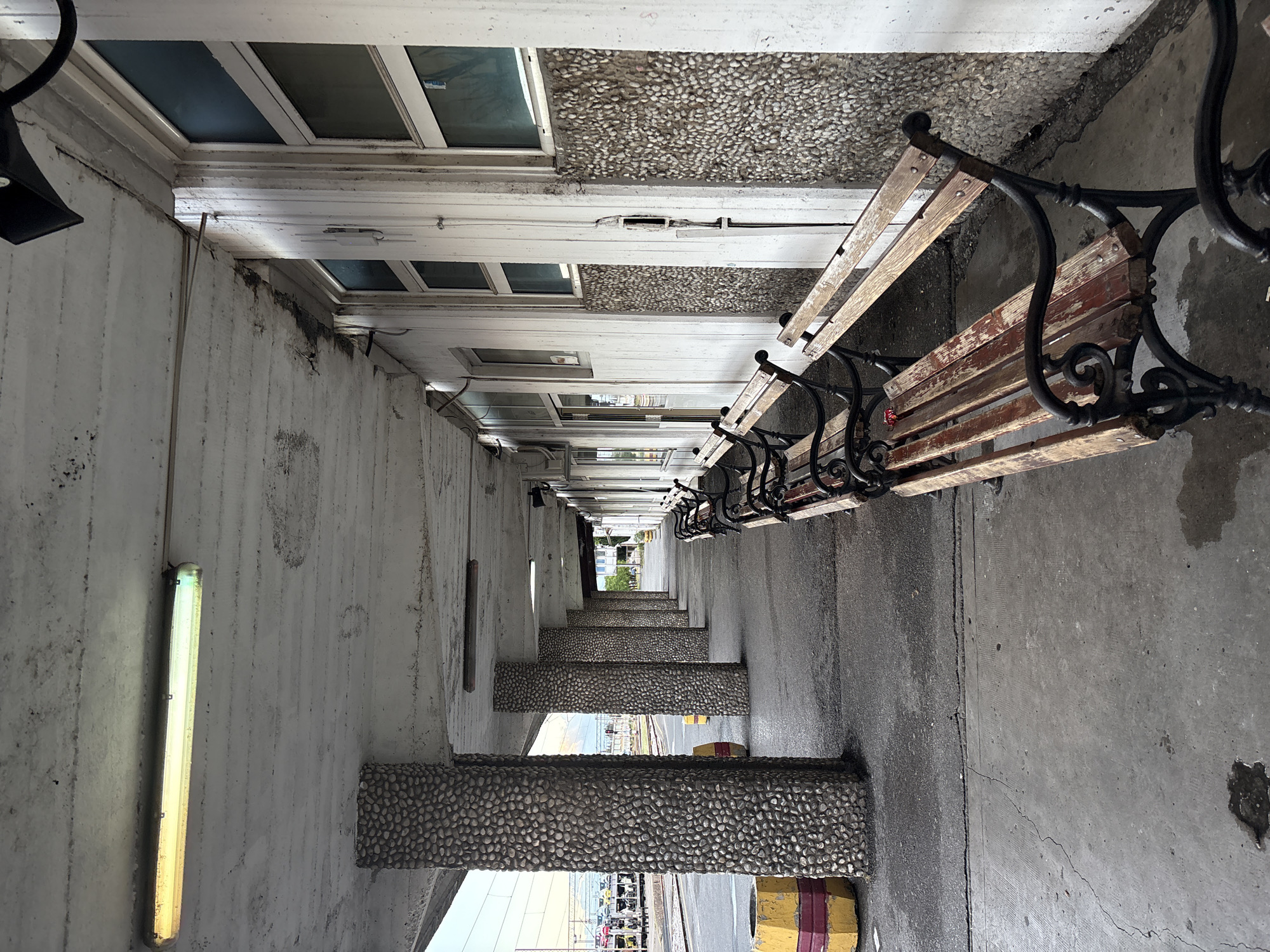 The image depicts a long, narrow pedestrian walkway or corridor in Podgorica, Montenegro. The space is defined by a concrete wall on the left and a row of benches on the right. The wall has rectangular lights and supports that are covered with small pebbles. Above the right side are a series of windows with white frames. Several benches line the right side, with black wrought iron armrests and weathered wooden slats. The concrete floor has cracks and patches. In the distance, the corridor opens up to a street scene with a visible parking area. The viewpoint is along the corridor, causing the perspective to narrow in the distance.