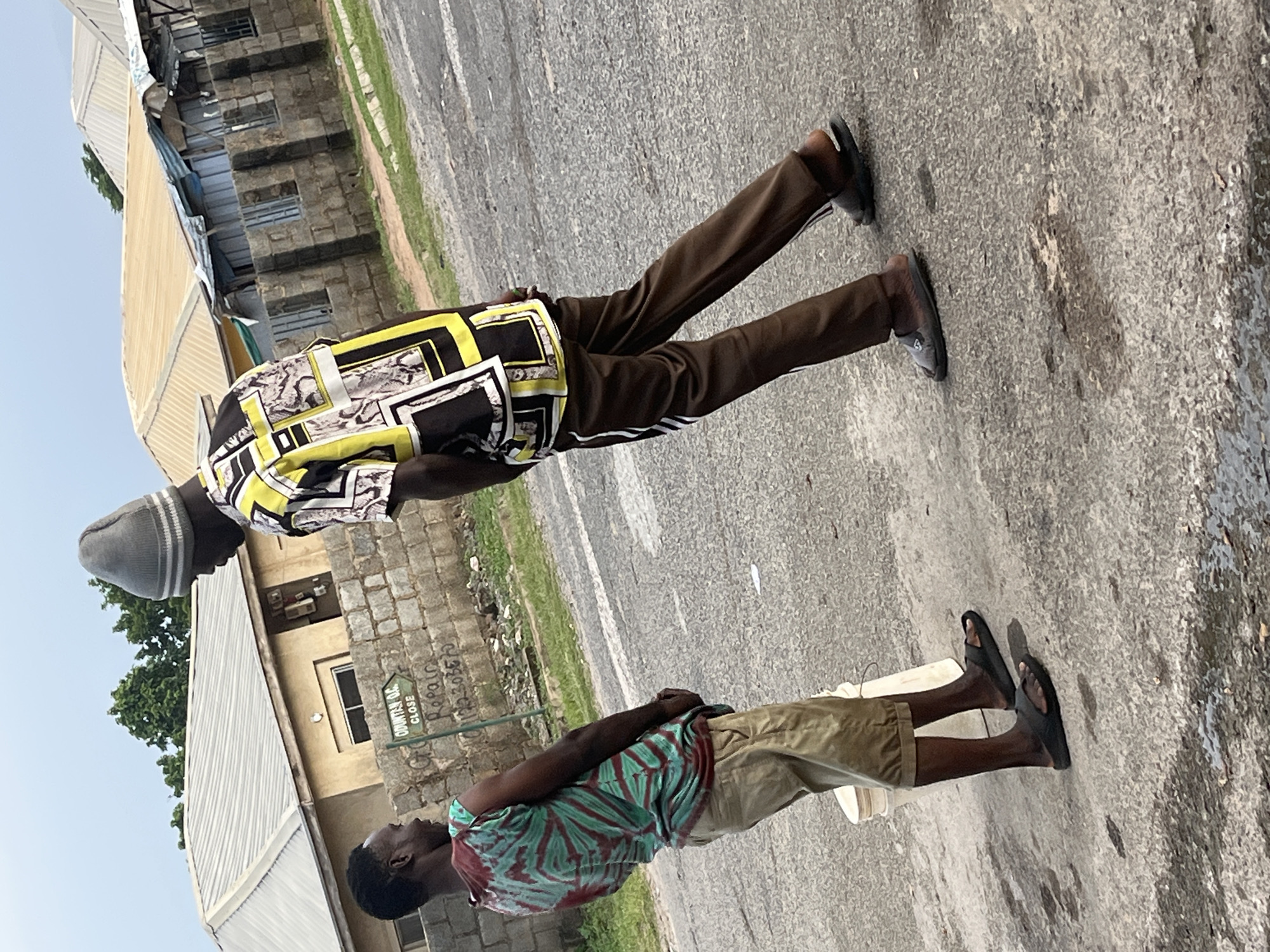 Two young men stand on a paved road in Abuja, Nigeria, engaged in conversation. The man on the left, wearing a tie-dye sleeveless shirt and khaki shorts, is facing the other man. He has dark skin and short black hair and is wearing black sandals. The man on the right, taller and wearing a grey beanie, a black, yellow, and white patterned short-sleeved shirt, and brown athletic pants, is turned slightly away from the viewer. He is also wearing dark sandals. Behind them, a low brick wall runs horizontally, with a green signpost pointing left reading "ODUNTAN O.C. CLOSE" and some graffiti. Beyond the wall, buildings with corrugated metal roofs are visible, suggesting a residential or commercial area. The road surface is grey asphalt, with some patches of wetness or staining. The sky is overcast, indicating a cloudy day. A white plastic bucket is positioned on the ground to the left of the man in the tie-dye shirt.