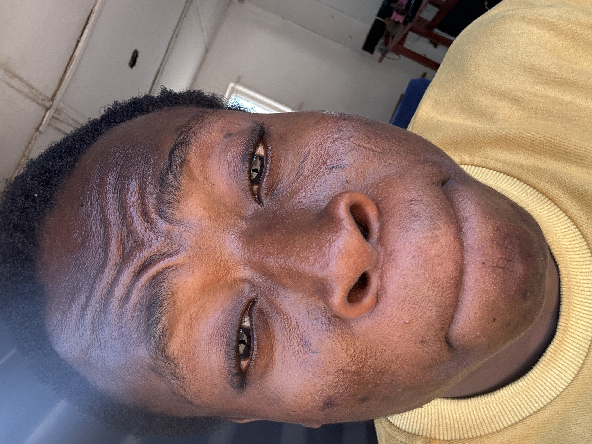 A close-up, rotated portrait captures a person with dark skin, short black hair, and brown eyes. The person is wearing a mustard-yellow, crew-neck sweater. The background features a partially visible white wall and ceiling, with a hint of a window and some furniture. The location is unidentifiable within the city of Unknown, Unknown.