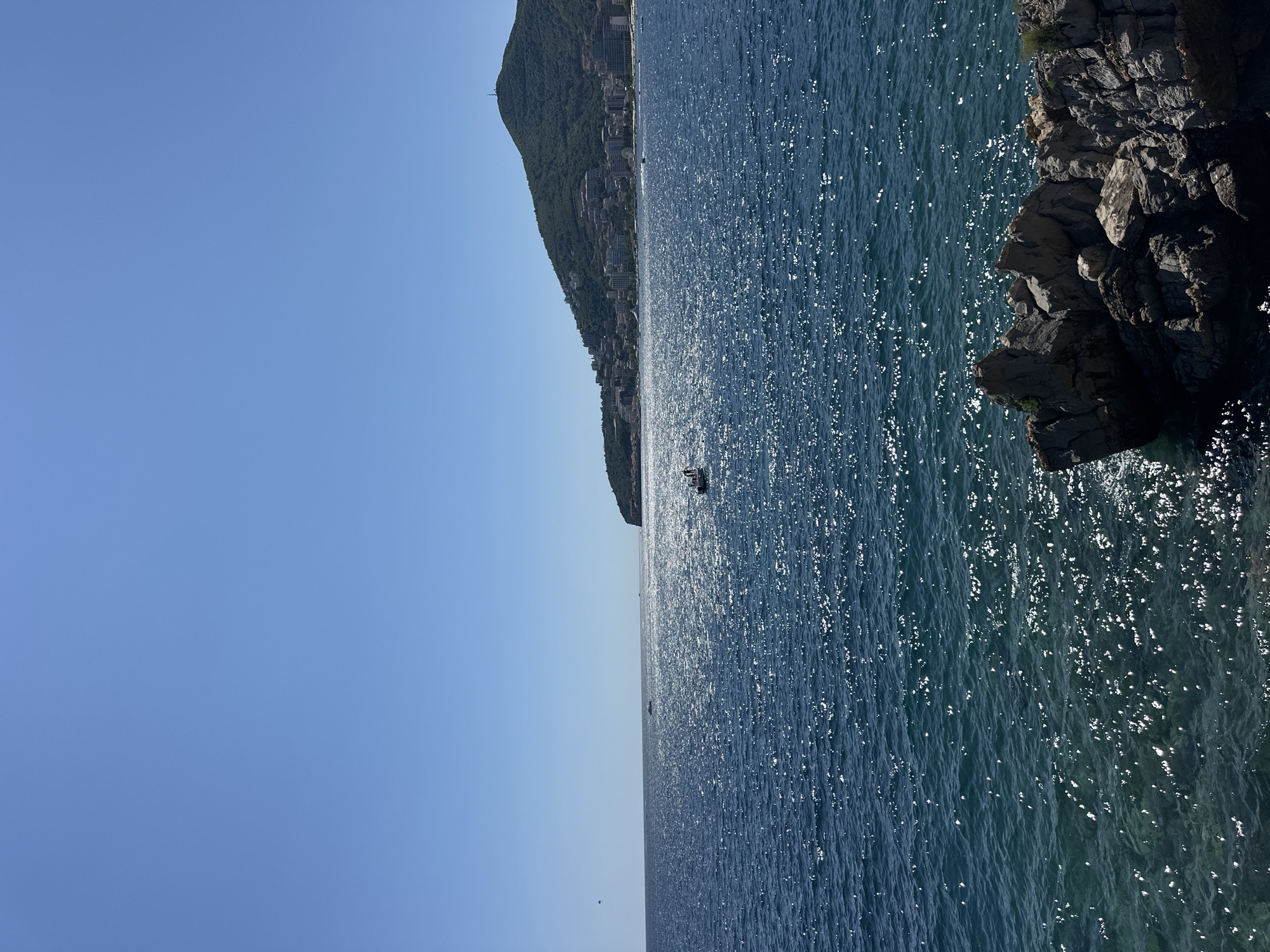A wide shot captures the Adriatic Sea under a clear, bright blue sky. In the foreground, rough, dark rocks jut out of the water on the right side. The sea itself is a deep blue, with sunlight glinting off its surface, creating a shimmering effect. Further out, a small white boat with at least two people onboard is visible in the middle distance. In the background, a green, tree-covered hill rises, dotted with buildings and structures that make up the city of Budva, Montenegro. The buildings are a mix of white and terracotta, cascading down the hillside towards the coast. The overall impression is one of a sunny, coastal day.
