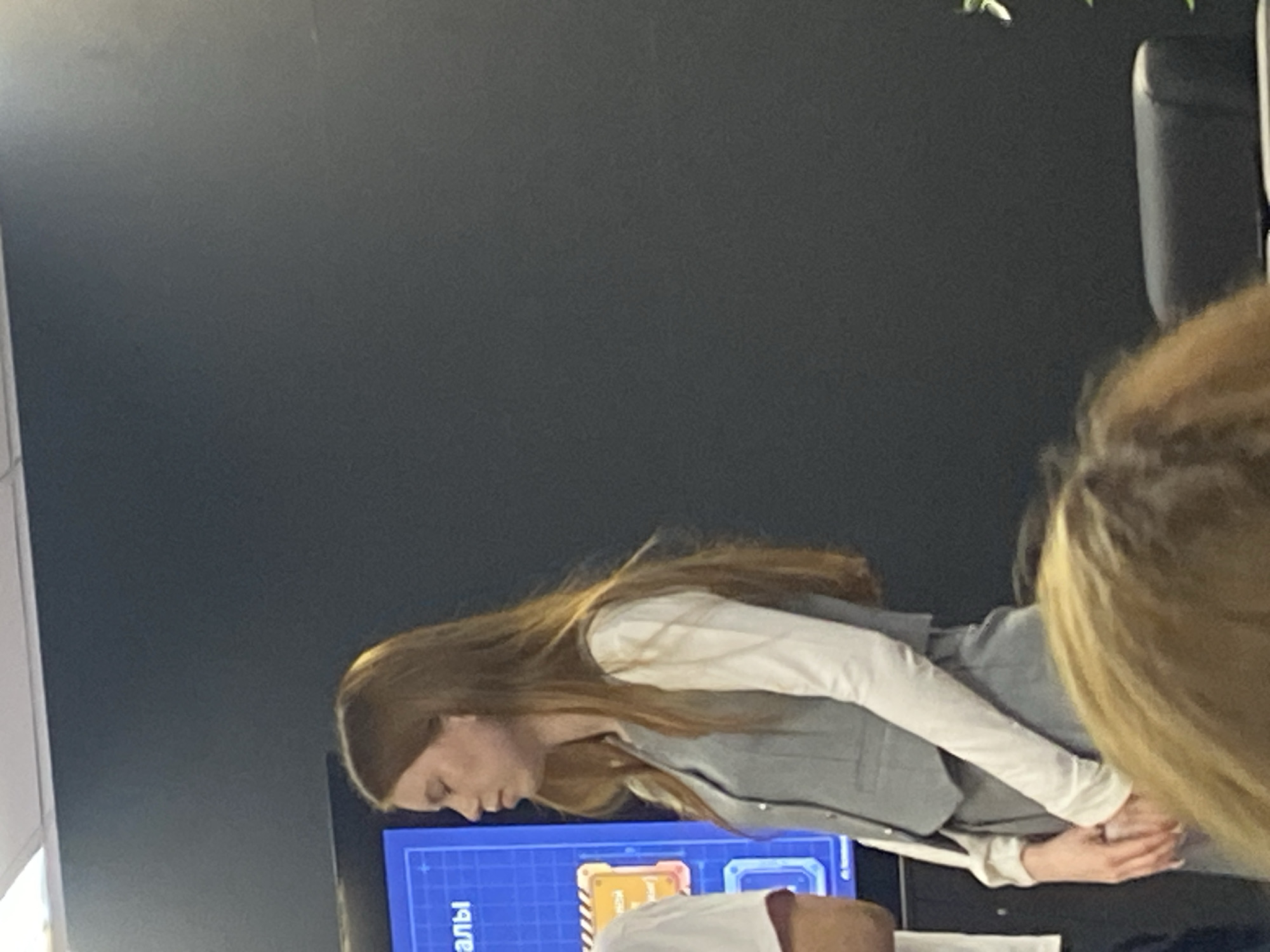 A young woman with long, auburn hair is standing in front of a dark gray wall and a blue screen, her head tilted down. She is wearing a white long-sleeved shirt and a gray vest and matching pants. Her hands are clasped in front of her. Another person's head and shoulder are visible in the foreground, out of focus, with blonde hair. The scene appears to be indoors, possibly an office or conference room, based on the presentation screen and the professional attire. The overall lighting is dim.