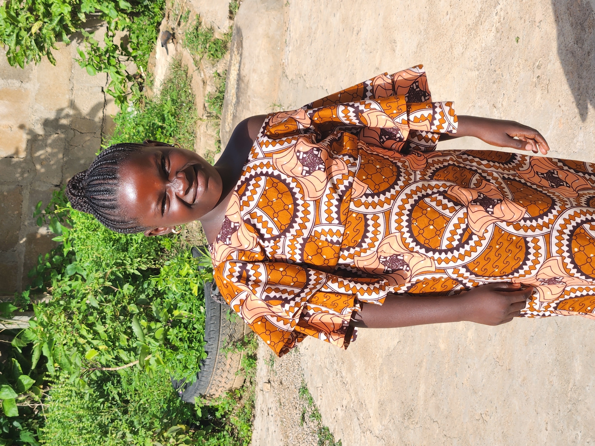 A young girl with braided hair stands outdoors, smiling at the camera. She is wearing a colorful, off-the-shoulder dress with ruffled sleeves, featuring a geometric pattern in shades of orange, white, and brown. The background consists of lush green vegetation, including plants in a discarded tire, and a textured brick wall. The ground is a rough, sandy or concrete surface, and a shadow is cast on the right side of the frame. The overall setting appears to be a residential area in Sabon Tasha, Nigeria.