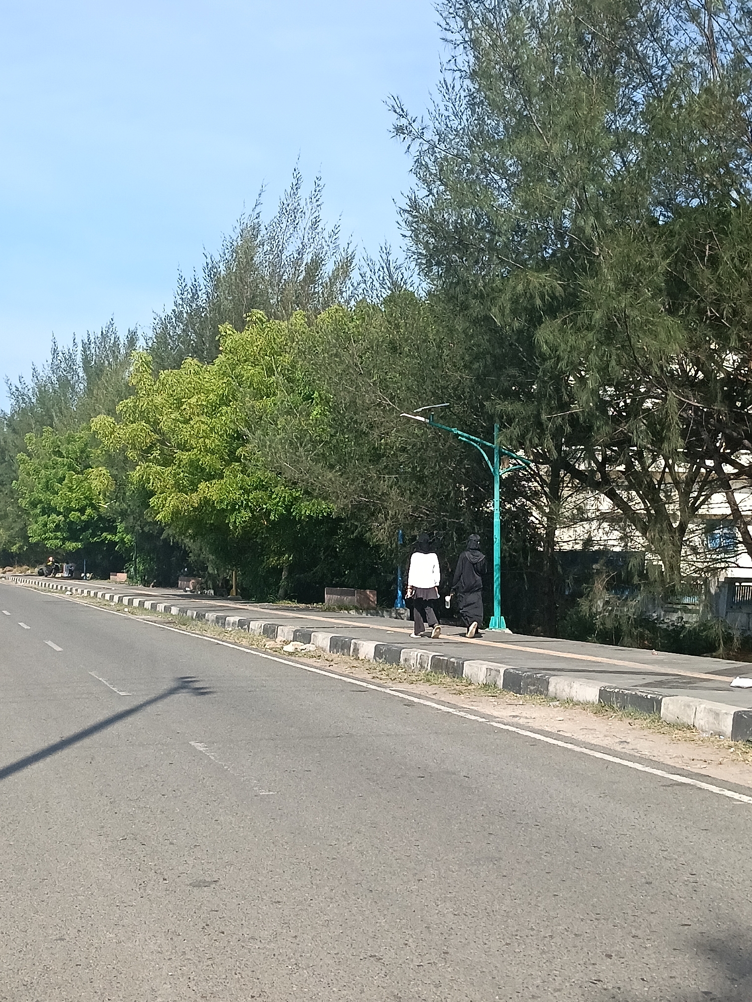 This image captures a daytime scene in Banda Aceh, Indonesia, likely along a coastal road or park. In the foreground, a paved road with white lane markings stretches into the distance. To the right of the road, a sidewalk runs parallel, bordered by a curb with alternating black and white markings. A teal-colored street lamp stands on the sidewalk. Two individuals are walking away from the camera on the sidewalk. The person on the left wears a white jacket and black pants, while the person on the right is dressed in a black hijab and robe. Behind the sidewalk, a dense line of trees, including tall, needle-leafed varieties and lush green deciduous trees, forms a natural barrier. In the background, a white building with windows is partially visible through the foliage. The sky above is clear and blue. The overall impression is of a well-maintained public space with natural elements.