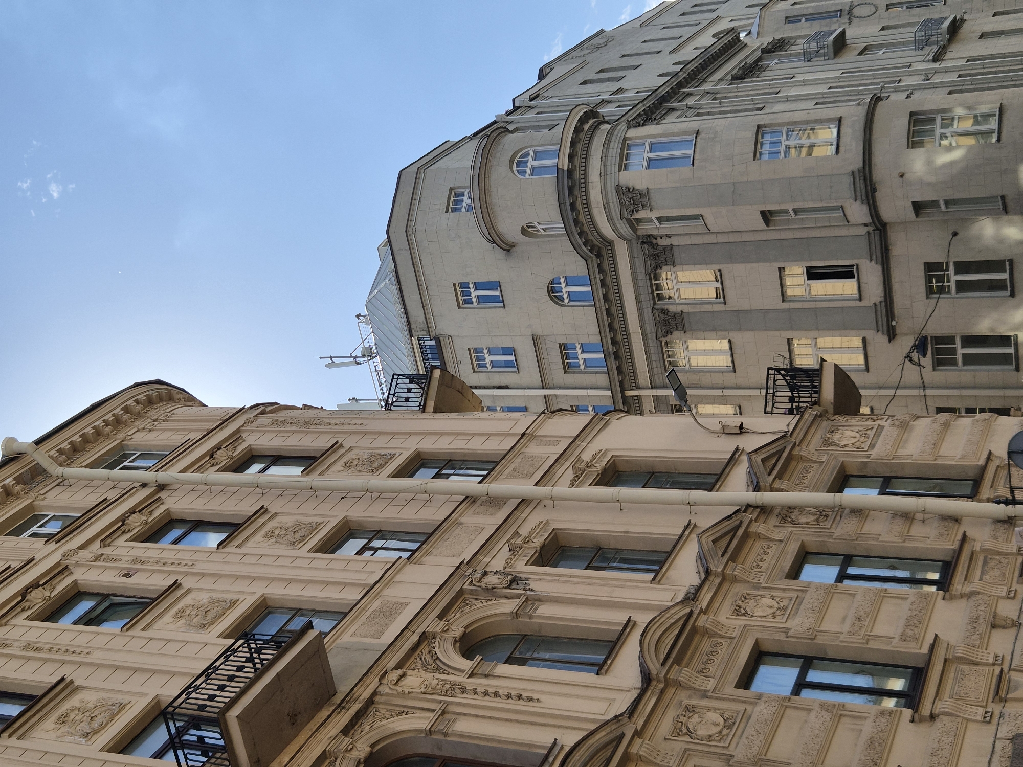 This image depicts a street-level view of ornate, European-style buildings against a clear blue sky. The building in the foreground, on the left, is a light beige color with detailed architectural elements, including decorative carvings above the windows and arched windows on the lower levels. It features black wrought-iron balconies and a light-colored downspout running vertically along its facade. To the right, a second building, lighter gray in color, curves around a corner. This building also has multiple windows, some arched and some rectangular, and features a rounded bay window section. Both buildings exhibit multiple stories and appear to be residential or commercial structures. The lighting suggests it is daytime with the sun partially obscured, casting shadows. The scene is devoid of people. The architectural style is consistent with older European city centers.