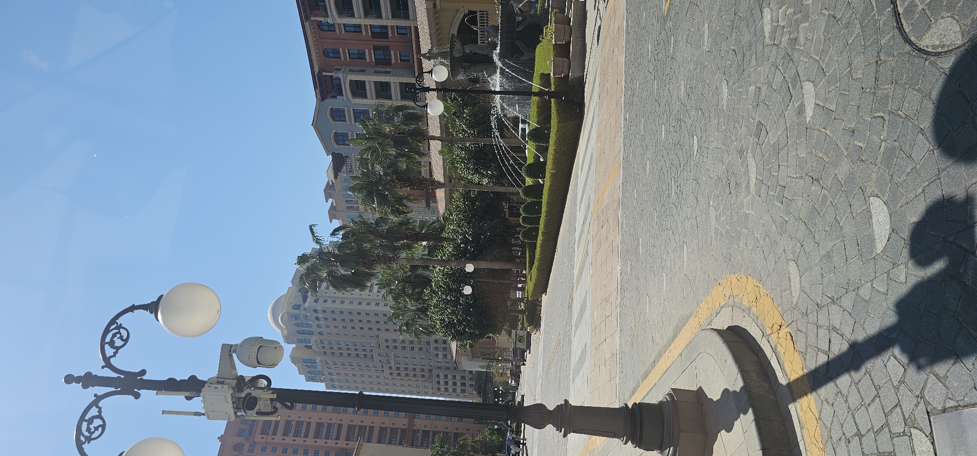 The image depicts a street scene in Doha, Qatar, specifically within the Pearl-Qatar development, known for its Venetian-inspired architecture and luxurious residences. A lamppost with two globe lights and attached security cameras stands prominently in the foreground on the left. The street is paved with cobblestones, and a yellow line marks the edge of the pavement. In the background, several tall buildings with a distinct architectural style, featuring curved balconies and beige facades, rise against a clear blue sky. Palm trees and manicured hedges are visible, adding greenery to the urban landscape. A fountain with water spraying upwards is located on the right side of the image. The scene appears to be a public area, possibly a promenade or a residential street within a resort-like setting. There are no people actively engaged in any specific action, though some cars are parked or driving in the distance. The overall impression is one of a well-maintained, upscale urban environment characteristic of modern developments in Doha.