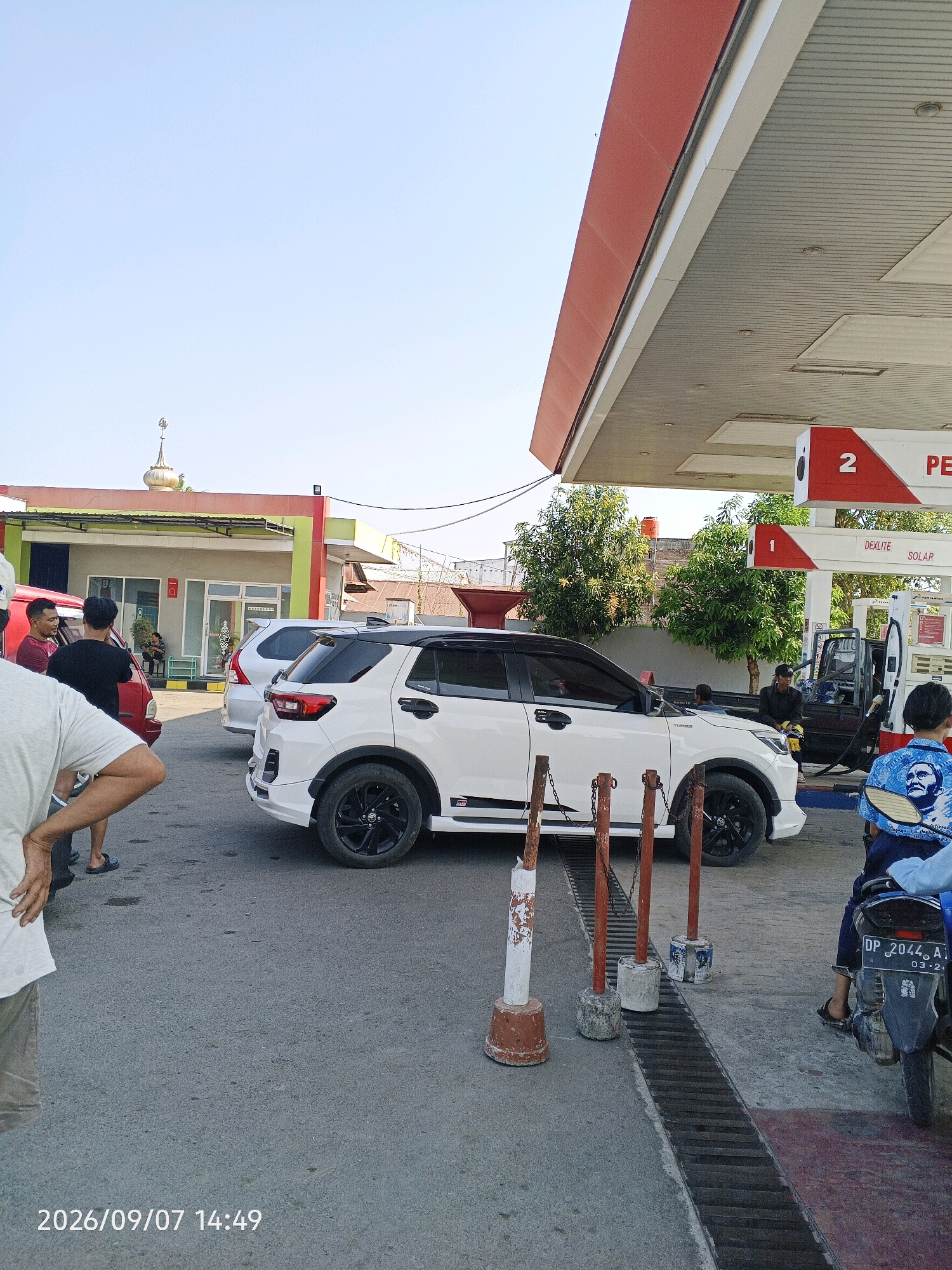The image depicts a daytime scene at a Pertamina gas station in Allakuang, Indonesia. The sky is clear and blue. A white SUV is parked at a gas pump under the red and white awning of the station. A man is visible near the pump, possibly refueling the vehicle. In the foreground, several people are standing and interacting, with a red car partially visible on the left. A motorcycle with a rider wearing a blue shirt with a portrait on it is parked to the right, facing away from the camera. The ground is paved, and a drainage grate runs along the edge of the pump area. In the background, a building with a green and red facade and a domed structure, possibly a mosque, is visible. The overall atmosphere suggests a routine stop at a local gas station.