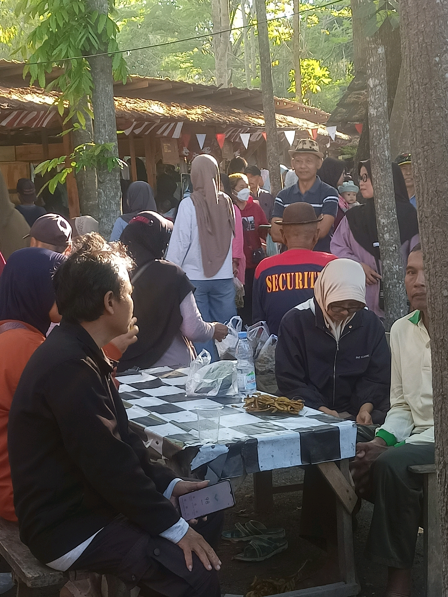 In Karungan, Indonesia, an outdoor gathering takes place near a rustic building with a thatched roof adorned with red and white pennants. Sunlight filters through the trees, illuminating a group of people. In the foreground, a man in a black jacket sits at a table covered with a checkered cloth, holding a smartphone. Beside him, another person in a dark jacket and headscarf is seated, looking down. Behind them, a man in a blue shirt and a hat is visible, as is a person wearing a jacket with "SECURITY" printed in orange on the back. Other individuals, some wearing headscarves, are also present, contributing to the lively atmosphere. The ground beneath the table is strewn with a few pairs of sandals.