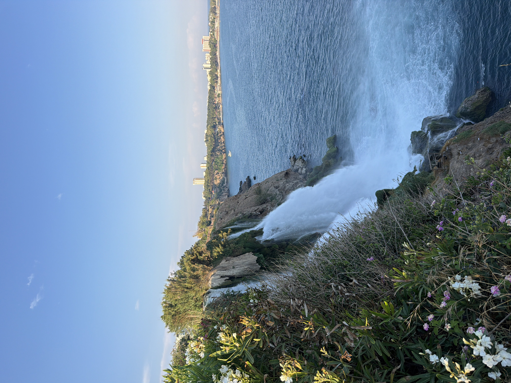 This image captures the Duden Waterfalls in Antalya, Turkey. The scene is dominated by a cascading waterfall that plunges from a cliff face into the Mediterranean Sea. Lush green vegetation, including flowering bushes with white and purple blossoms, borders the top of the cliff. The sea stretches out to the horizon, with a city skyline visible in the distance, characterized by tall buildings and trees. The sky is a clear blue with a few scattered clouds. The perspective is from the cliff edge, looking down and out towards the sea. There are no people visible in the image.