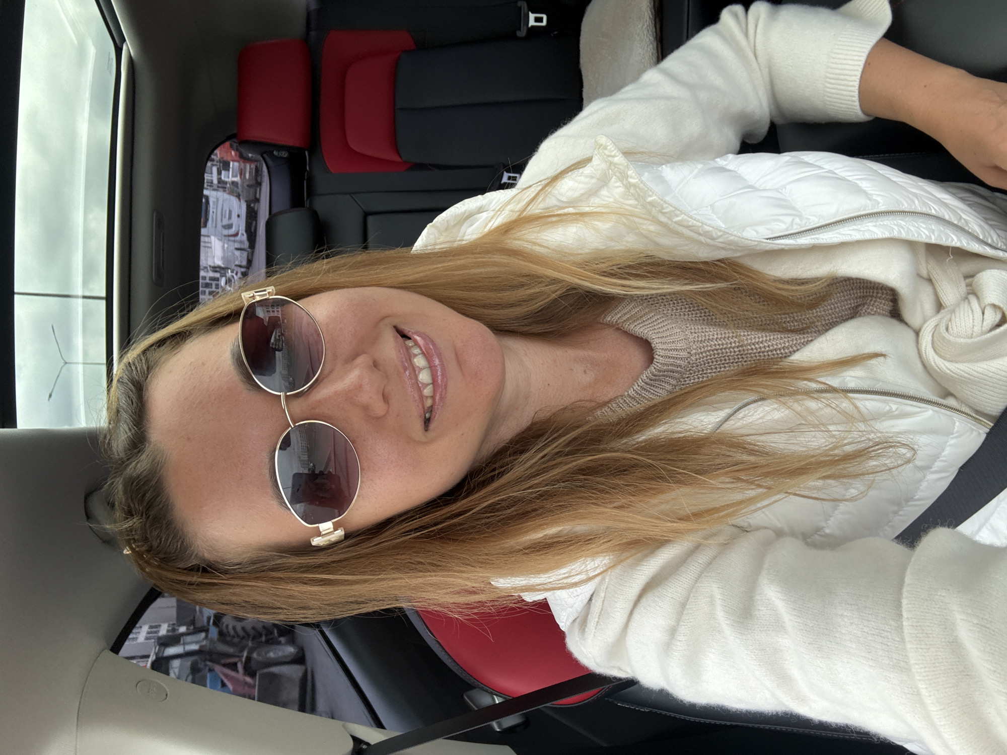 A woman with long, blonde hair, wearing sunglasses and a white puffer vest over a beige sweater, is taking a selfie in the passenger seat of a car. The car's interior features red and black leather seats and a panoramic sunroof. Through the sunroof and windows, several trucks and a construction vehicle are visible, suggesting the car is on a road or in a commercial area. The background appears to be an overcast sky.