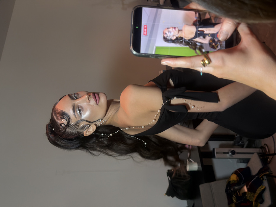 An indoor scene in the city of Paris, France, captures a styled female subject posing for a photograph being taken by a partially visible individual. The primary subject is a young adult female with dark hair styled in a high ponytail, featuring sculpted waves adorned with small white pearls on the forehead and temples. Her makeup includes prominent white eyeshadow and highlighted skin, with pink-toned lips. She wears a black off-the-shoulder dress with bow details on the sleeves. A string of small white pearls drapes from her left ear, extends across her back, and trails down her left arm, with additional individual pearls applied to her skin on the arm. She is positioned with her body turned to the right, looking back over her left shoulder directly towards the camera.
In the immediate foreground, the right hand of a second individual is visible, holding a vertical smartphone. The smartphone's screen displays a live preview of the subject, with a red "00:00:15" timer indicating a video recording is in progress. The individual holding the phone wears a gold-toned ring, possibly shaped like a snake, and a thin turquoise ring. The background consists of a plain light-colored wall. To the left, a dark table holds a black bag and a white mug. Beneath the table, a light-colored chair is draped with patterned clothing, and a silver-colored device is partially visible on the floor. The environment suggests a backstage area, dressing room, or studio setting.