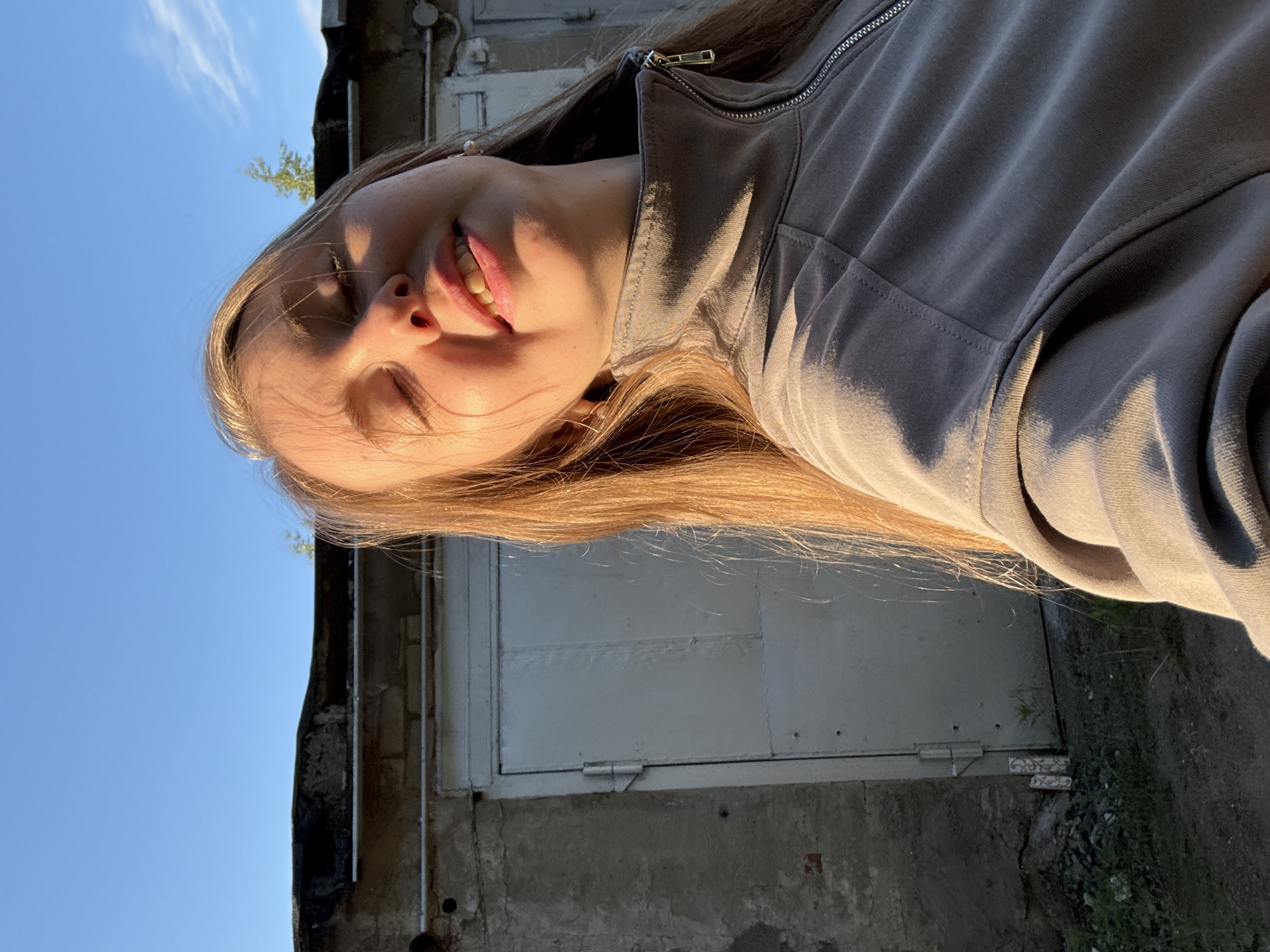 A low-angle, close-up portrait captures a young person, likely female, looking upwards with a broad smile and eyes mostly closed, possibly squinting from bright sunlight. Their long, light brown hair is illuminated by warm, golden light, framing their face and falling over their shoulders. They are wearing a grey or khaki-colored zippered hoodie, which also catches the sunlight, creating highlights and shadows.
The lighting suggests the photo was taken during golden hour, likely late afternoon or early evening. The sky is a clear, bright blue with a few wispy white clouds, forming a significant part of the background.
Behind the person, an outdoor setting is visible, including a weathered building or wall made of concrete and brick, partially covered by a light-colored panel or door. There is also some uneven ground with sparse grass and a small patch of green foliage from a distant tree or bush. The overall mood conveyed is one of happiness and enjoyment in natural light. There is no visible text in the image.