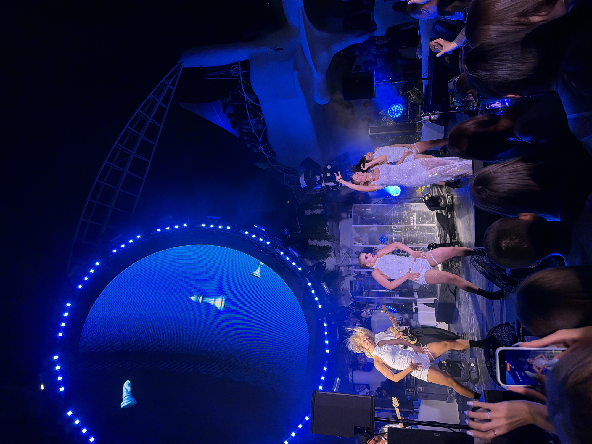 The image depicts a live performance by a musical artist on a stage at an outdoor venue in Odesa, Ukraine. A large, circular LED screen behind the performers displays abstract blue graphics, including what appear to be chess pieces. Three female performers are on stage, dressed in shimmering white outfits. The central performer is singing into a microphone, while the other two dance. They are accompanied by a live band, with a guitarist and bassist visible. The stage is lit with blue spotlights, and there are speakers and other equipment present. The audience is visible in the foreground, with some individuals holding up their phones to record the performance. In the background, a white architectural structure with a curved roof and railings is visible, suggesting an open-air amphitheater or similar venue. The overall atmosphere is energetic and festive, typical of a concert.