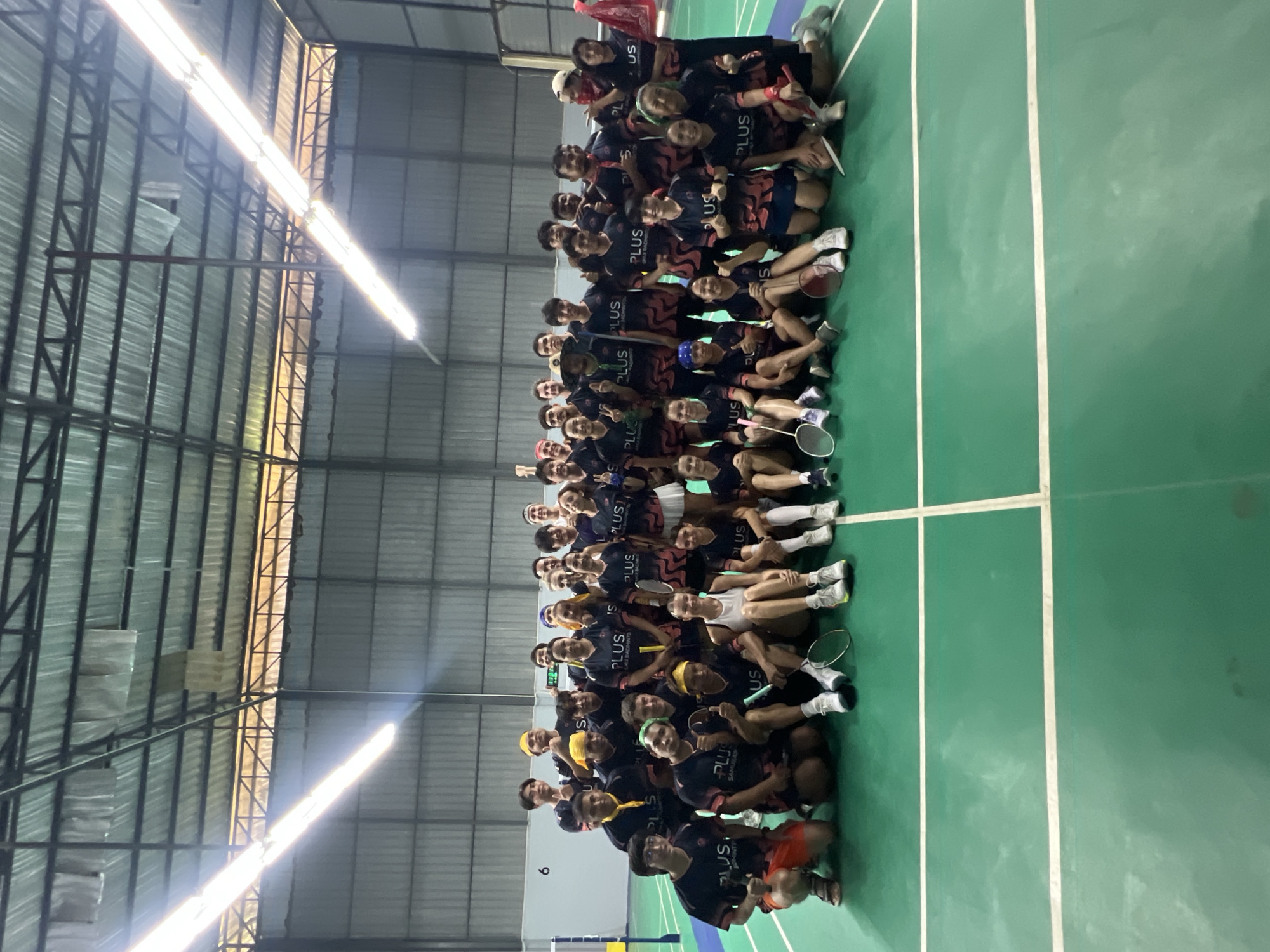 In the city of Ko Samui, Thailand, a group of approximately 30 individuals, comprising both males and females, are posing for a photograph on a green badminton court. They are all wearing matching dark blue t-shirts with pink accents and the text "PLUS" prominently displayed. Some individuals are also wearing headbands in various colors. The group appears to be a badminton team or club, as badminton rackets are visible in the hands of some members and on the court. The individuals are arranged in multiple rows, with some standing and others kneeling or sitting on the court. The environment is an indoor sports facility with a high ceiling featuring industrial lighting and corrugated metal walls. The court markings, including white lines, are clearly visible. The overall atmosphere suggests a team gathering or celebration.