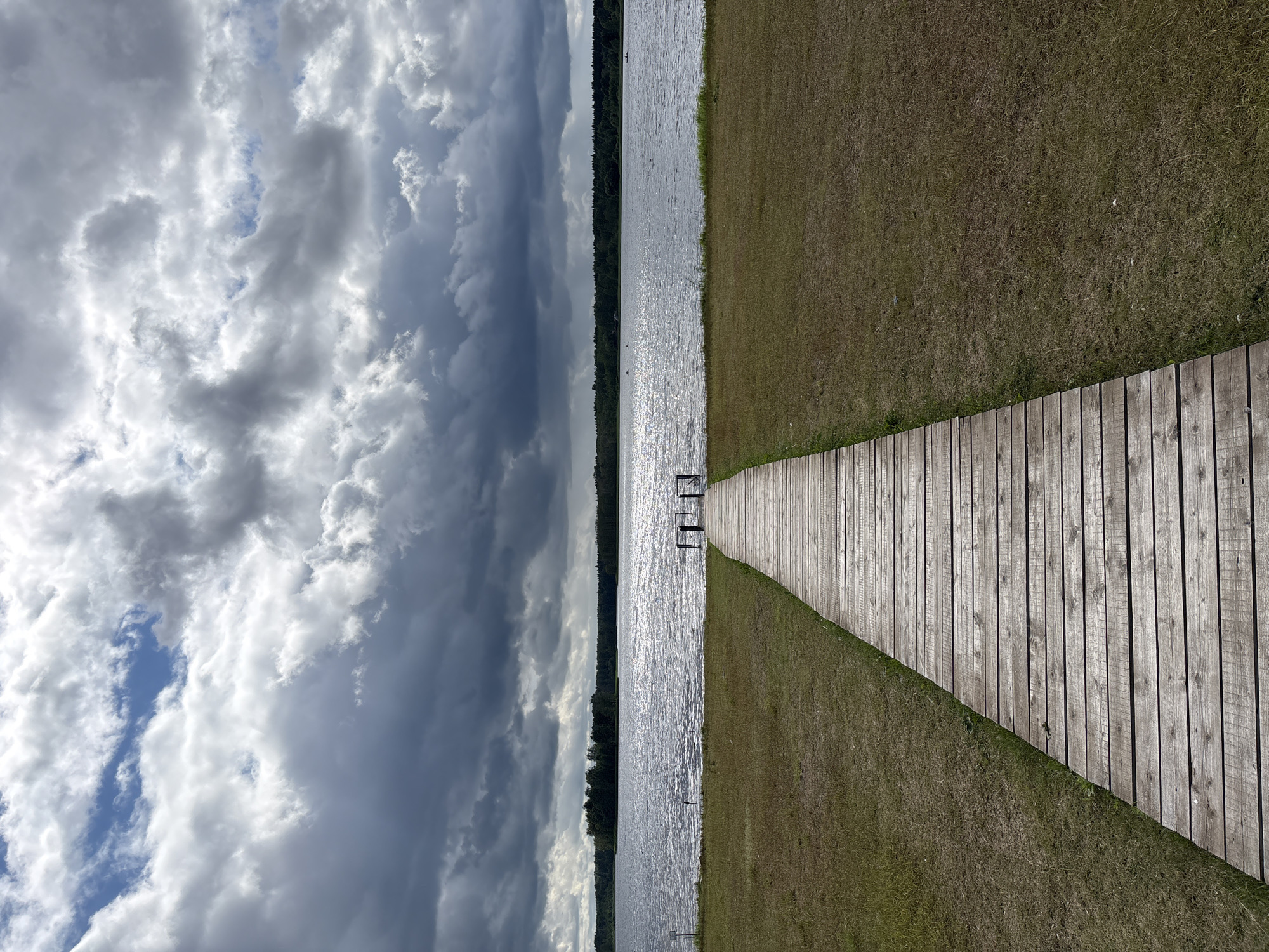 A wooden boardwalk extends from the foreground into a calm body of water, leading towards a distant tree line. The boardwalk is constructed from weathered wooden planks laid horizontally and appears to be situated on a grassy embankment. The body of water reflects the sky, which is dominated by dramatic, dark clouds interspersed with patches of brighter sky. The far bank of the water is lined with dense trees. The scene appears to be a natural landscape, possibly a lake or large river, with no visible buildings or urban structures.