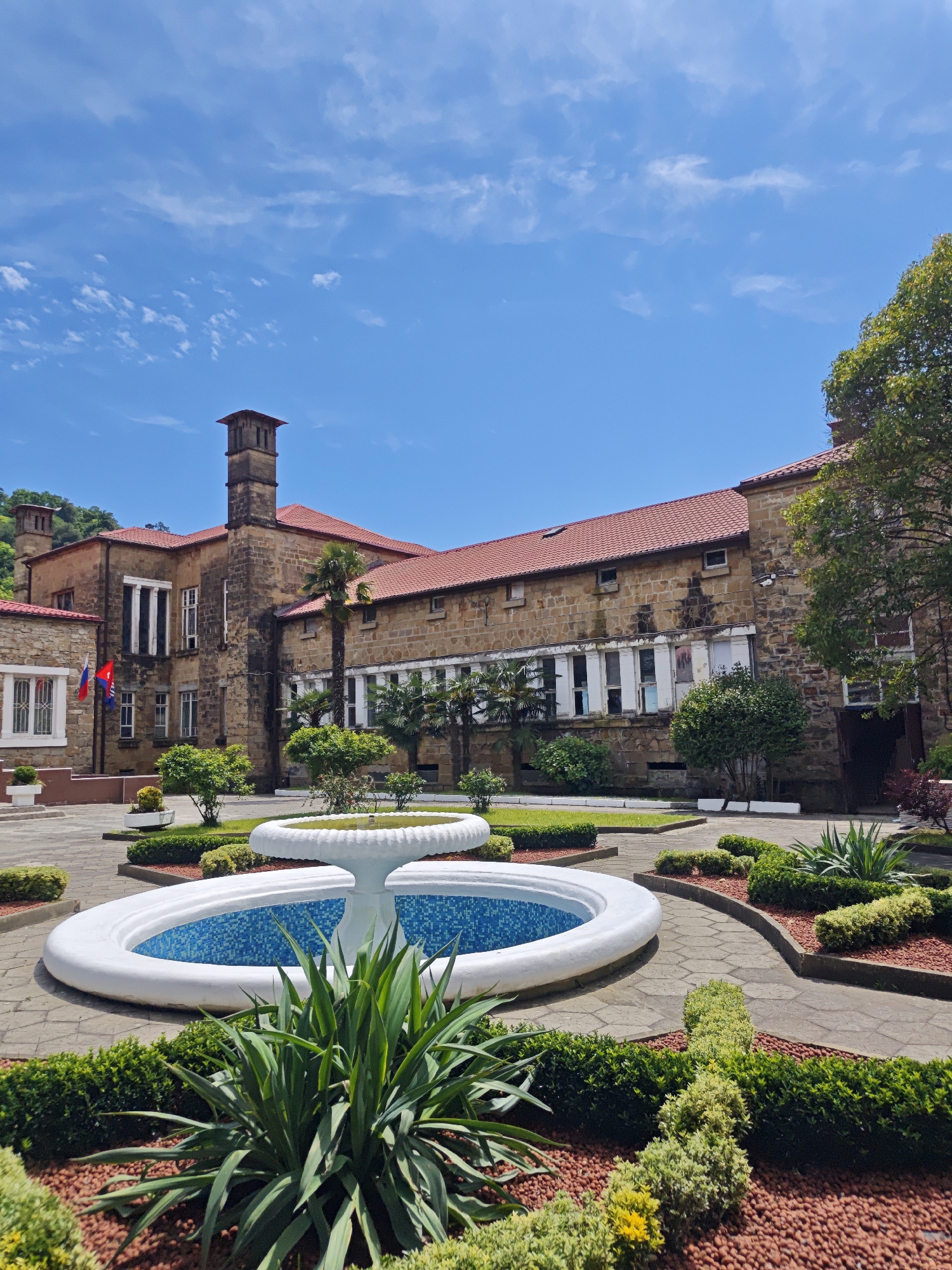 The image captures the architecture and landscape design of what appears to be the Villa Nadezhda in the city of Sochi, Russia. The main subject is a large, two-story building constructed of light-colored stone with a red tile roof. Its architecture features a tall chimney-like structure on the left side, a series of white-framed windows, and possible administrative flags. The building overlooks a courtyard with a fountain as its central feature. The fountain is circular with a white rim and a blue tiled interior. Manicured landscaping surrounds the fountain, including bushes, shrubs, and mulch. The environment suggests a sunny day with a clear blue sky and some scattered clouds. There are no people visible in the image.