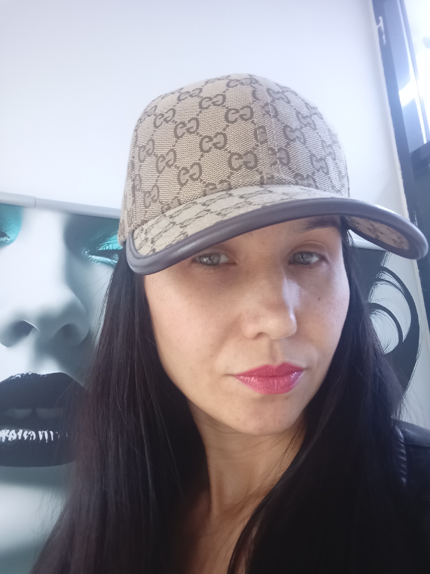 A woman with fair skin, dark hair, and pink lipstick is posing for a selfie. She is wearing a Gucci baseball cap. Behind her is a poster of a woman's face with heavy eye makeup and dark lipstick. The location is not identifiable as Izhevsk, Russia, based on the provided image.
